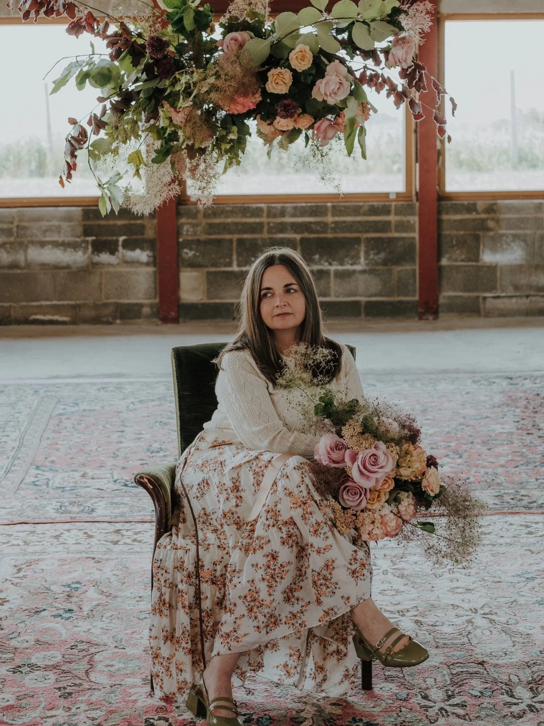 A woman with long dark hair, sitting on a vintage chair in a room with large windows and an exposed brick wall, holding a large bouquet of pink and cream flowers, and a hanging floral arrangement above her.
