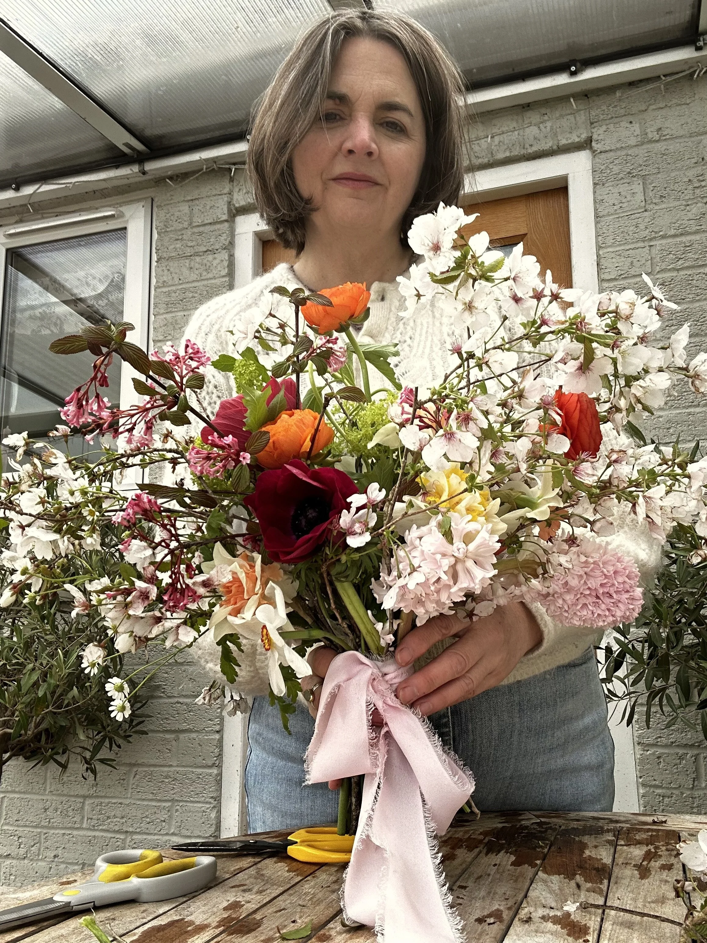 A woman holding a large bouquet of colorful flowers, including orange, red, pink, and white blossoms, while standing outdoors near a brick wall. She is wearing a cream-colored sweater and blue jeans, with flower scissors and a pink ribbon on the wood