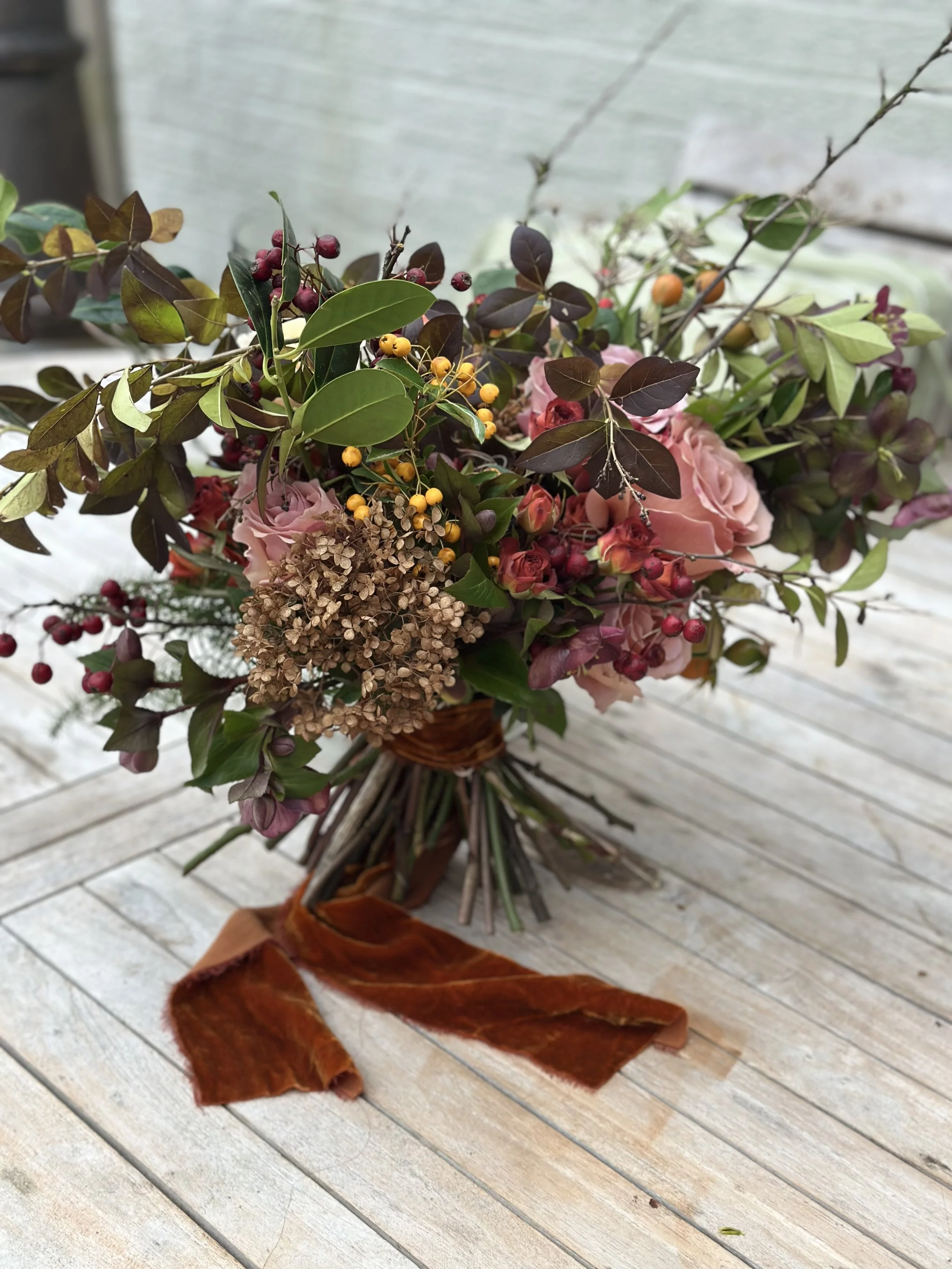 A floral bouquet with pink roses, yellow berries, brown dried flowers, green and burgundy leaves, and dried brown leaves on a white wooden surface.