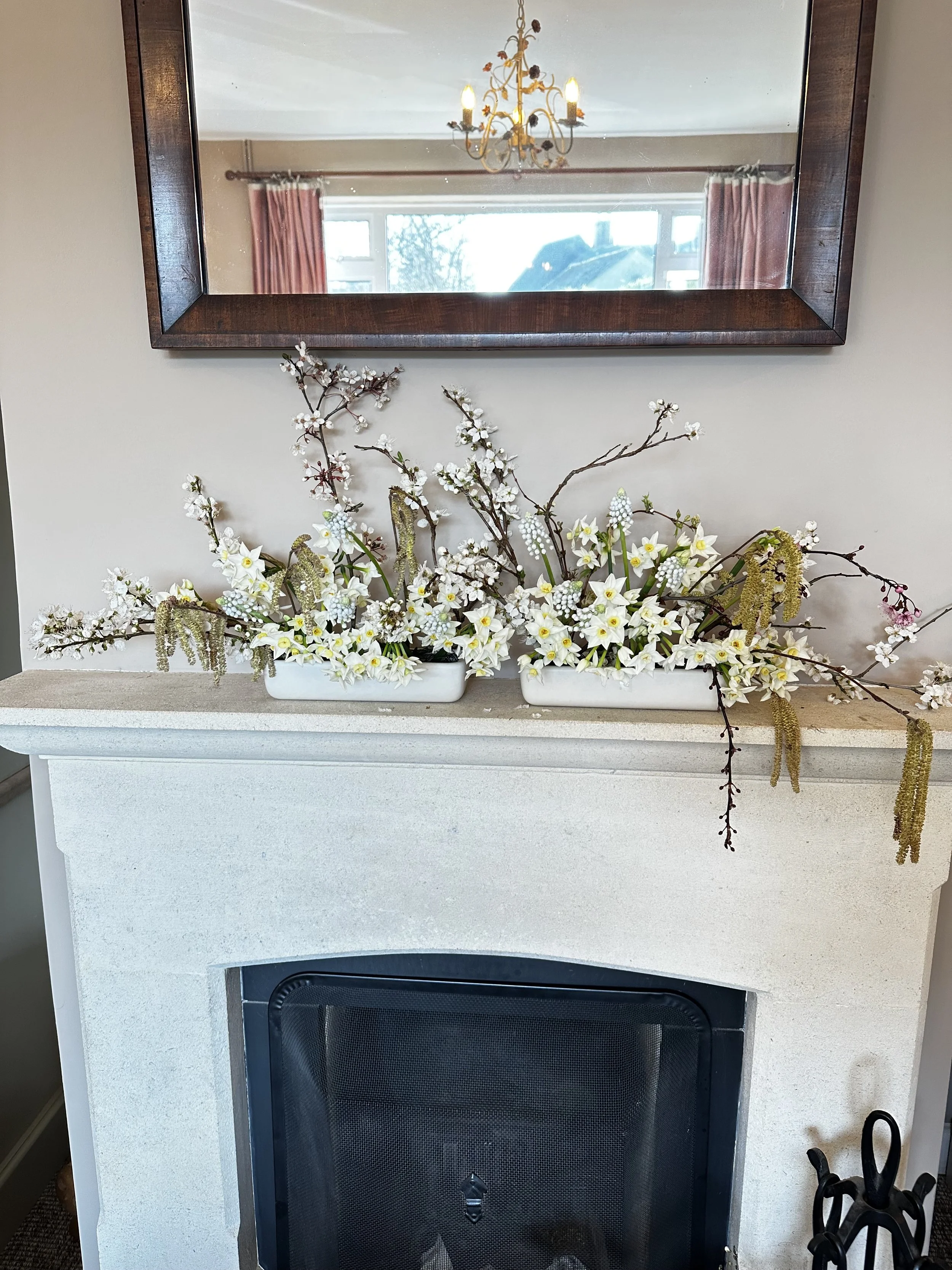 A fireplace with a decorative floral arrangement in front of it, a mirror above, and a chandelier reflected in the mirror.