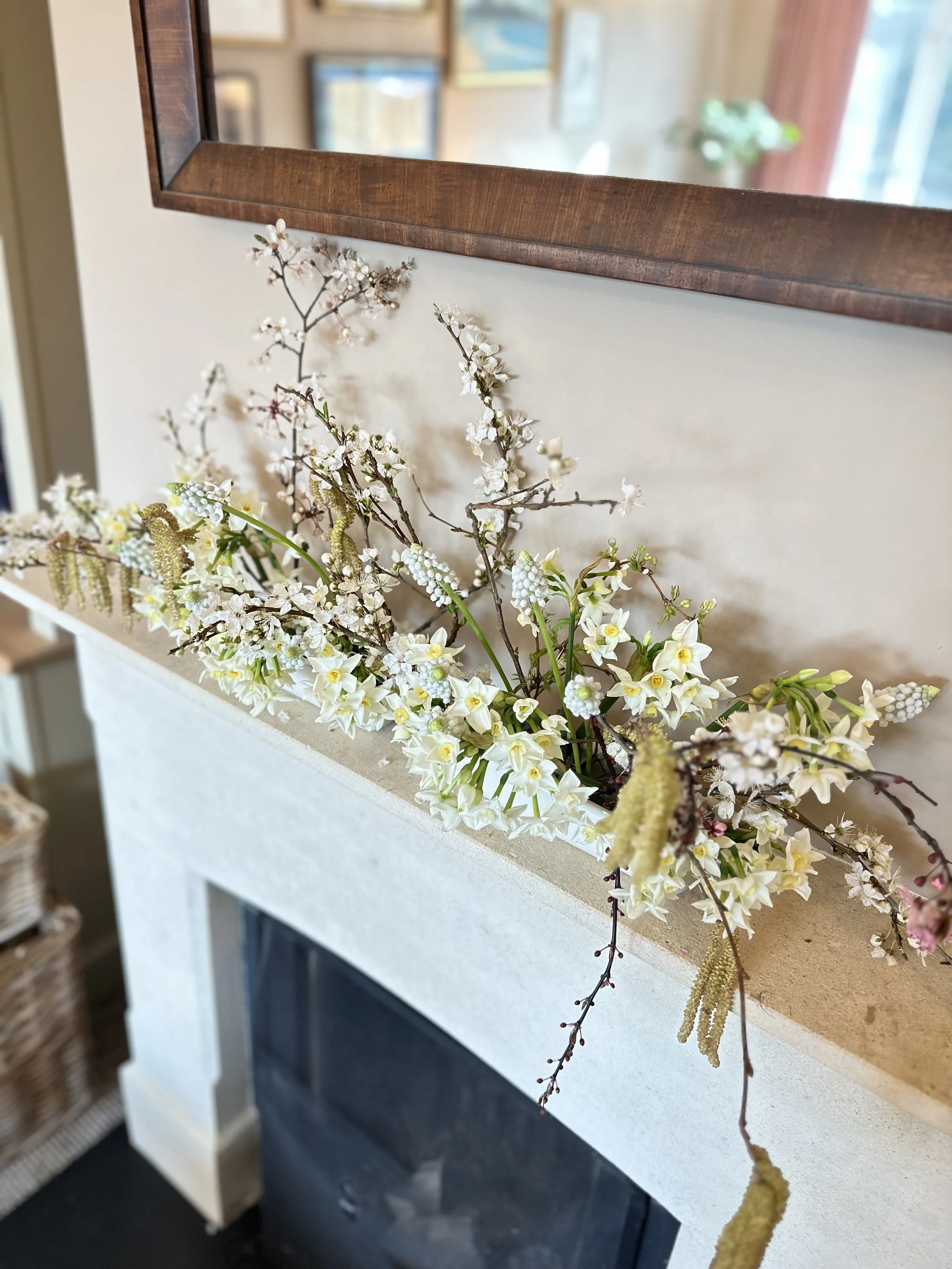 A close-up of a floral arrangement with white, pink, and yellow flowers and brown twigs in a white container, placed on a white surface in front of a mirror with a wooden frame. The mirror reflects a cozy interior with wall art and a window with curt