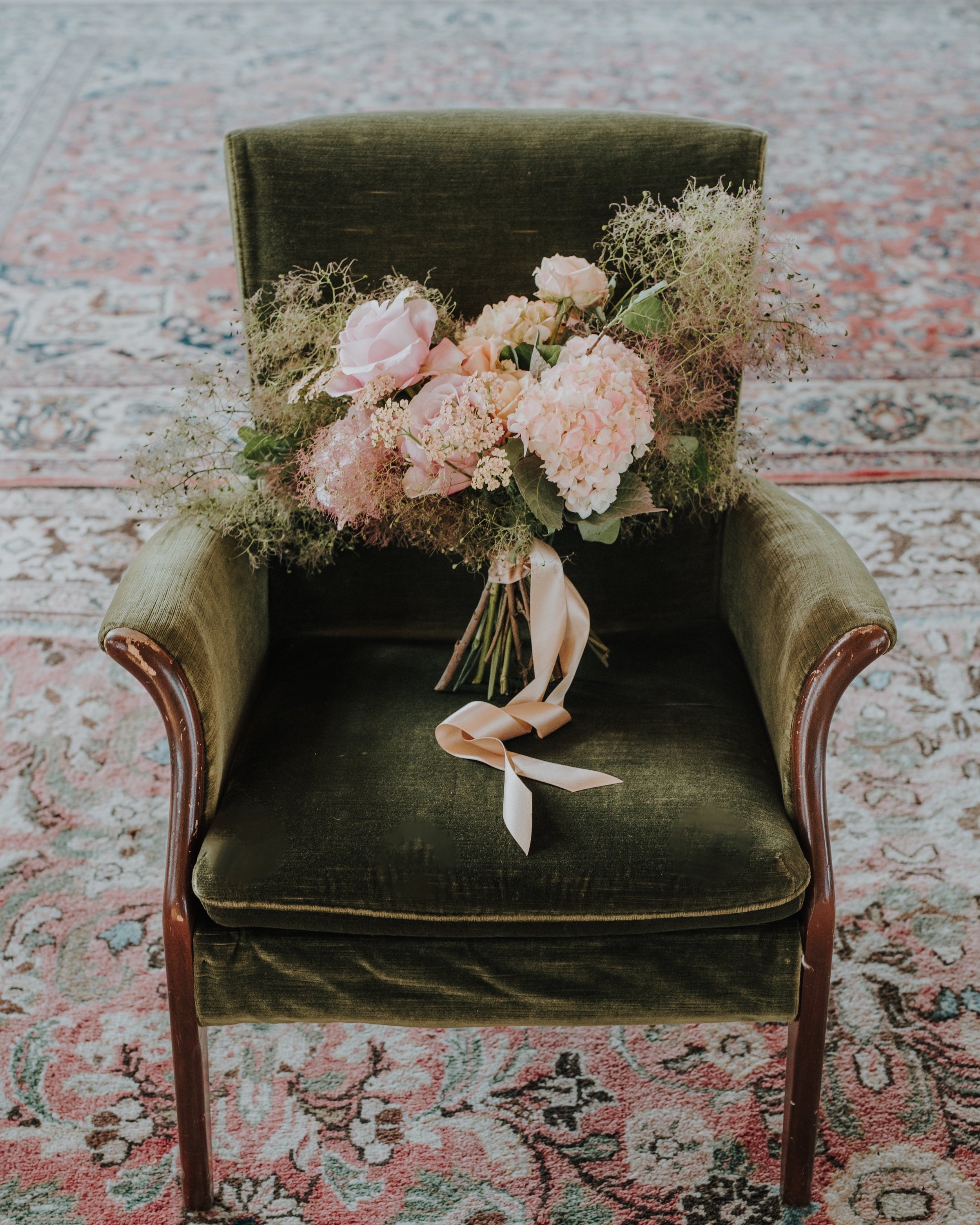 A vintage green velvet armchair with wooden arms, placed on a patterned pink and cream rug. Resting on the chair's back is a bouquet of pink and white flowers with greenery, tied with a beige ribbon.