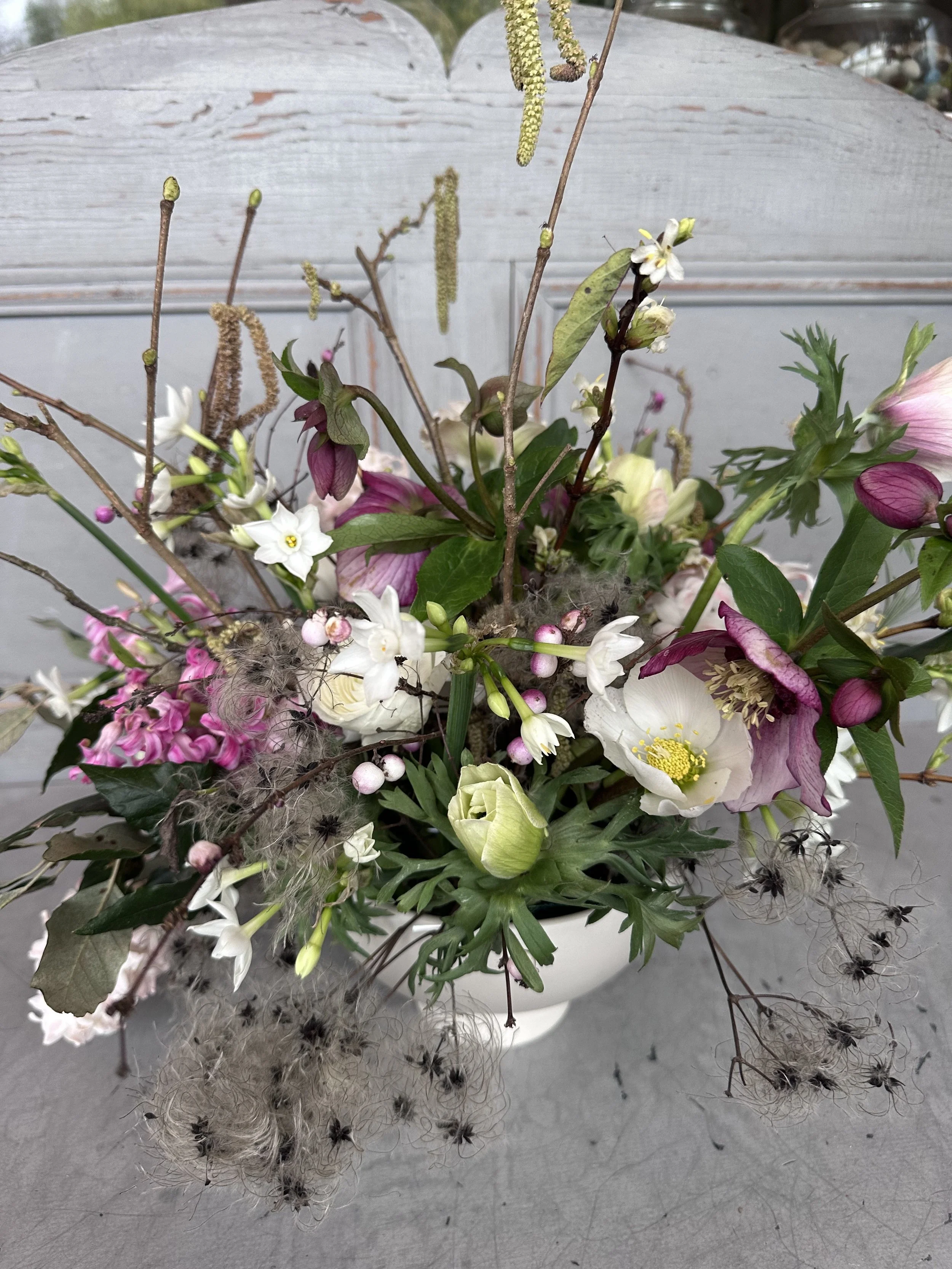 A bouquet of assorted flowers and foliage, including white, pink, and purple blooms, with some flower stems and dried branches, set against a gray background with a weathered white wooden surface.