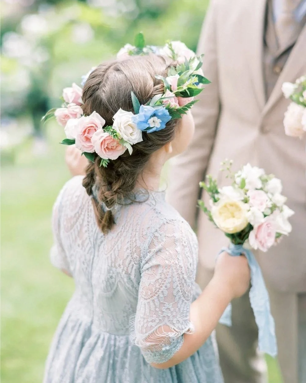 A young girl with braided hair wearing a lace dress and a floral crown, holding a small bouquet of flowers, standing outdoors during a wedding or similar event.