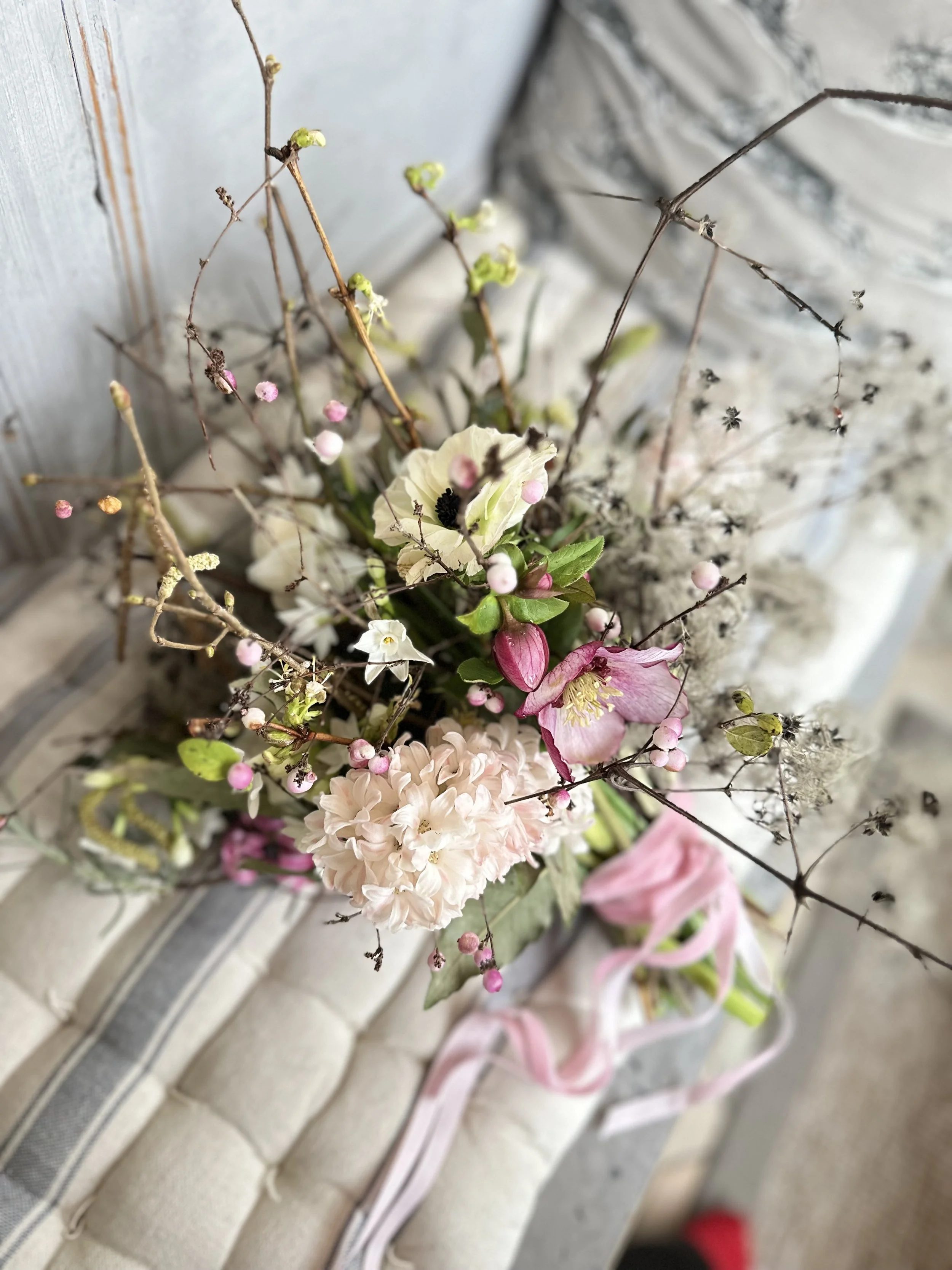 Close-up of a bouquet with various flowers and branches, placed on a textured white surface with a pink ribbon.