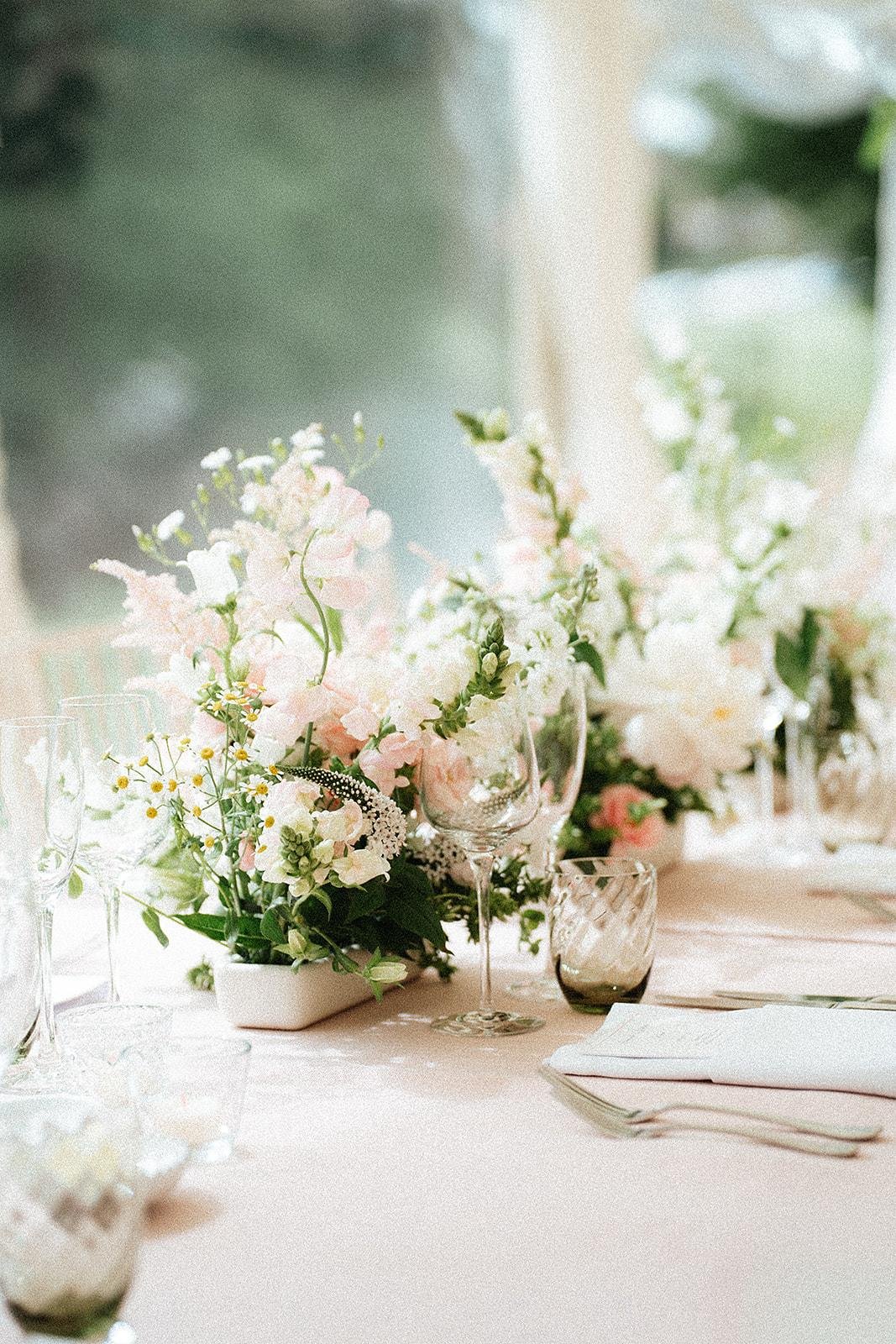 Elegant table setting with a floral centerpiece featuring pastel pink and white flowers, wine glasses, and tableware on a soft pink tablecloth.