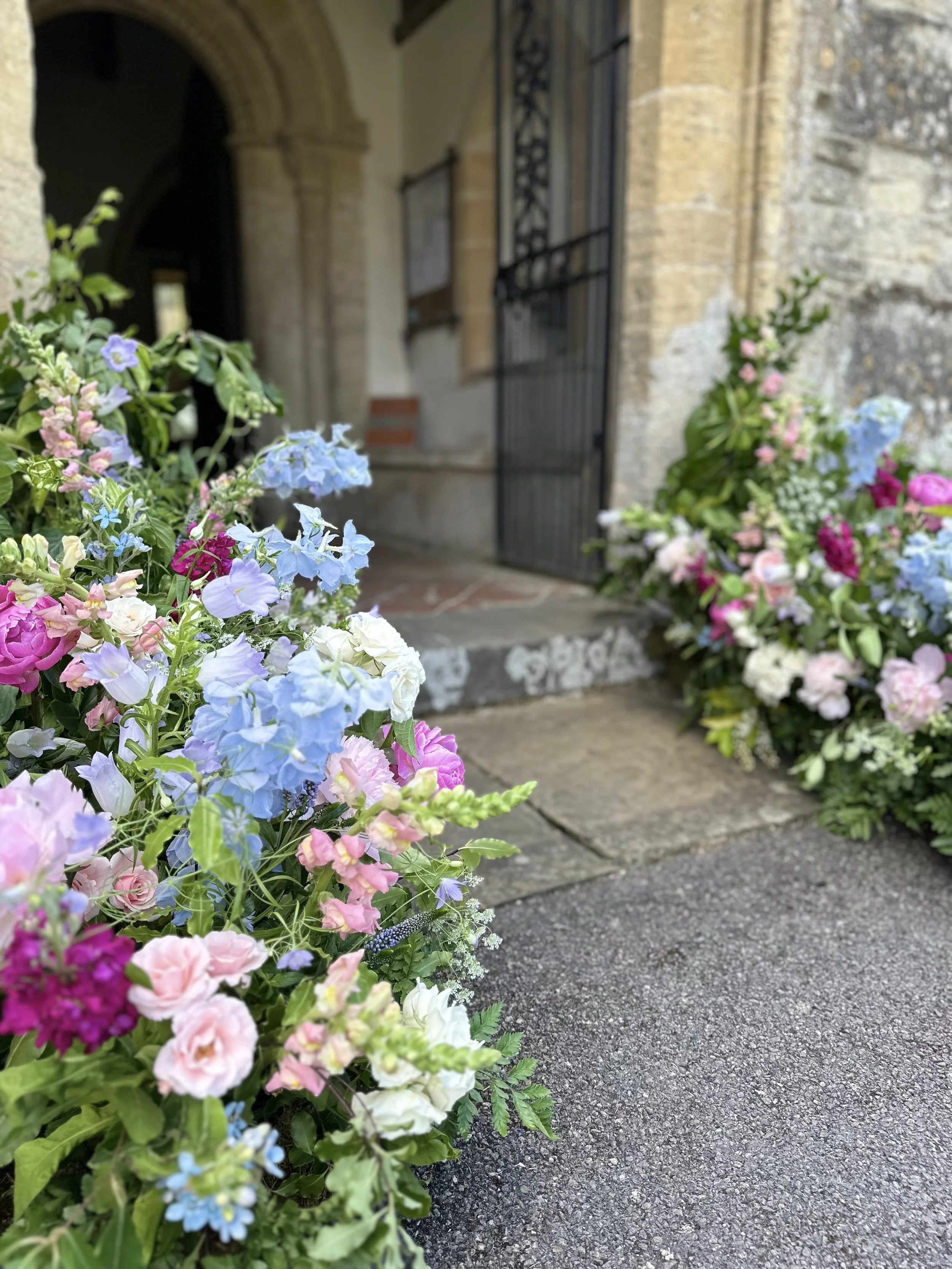 Colorful flower arrangement near the entrance of a stone building with arched doorway and open metal gate.