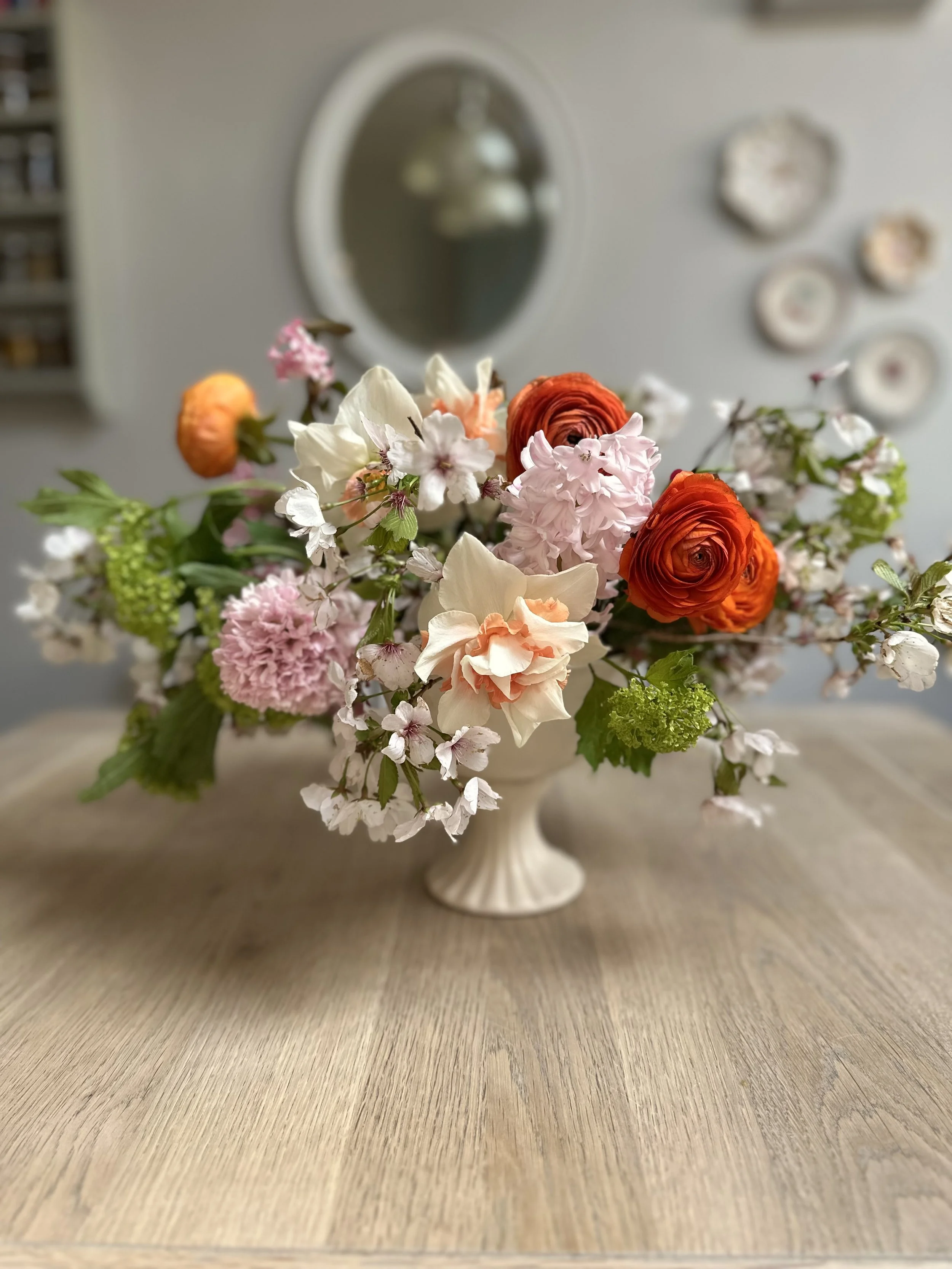 A white vase filled with a colorful bouquet of pink, white, orange, and green flowers placed on a wooden surface.