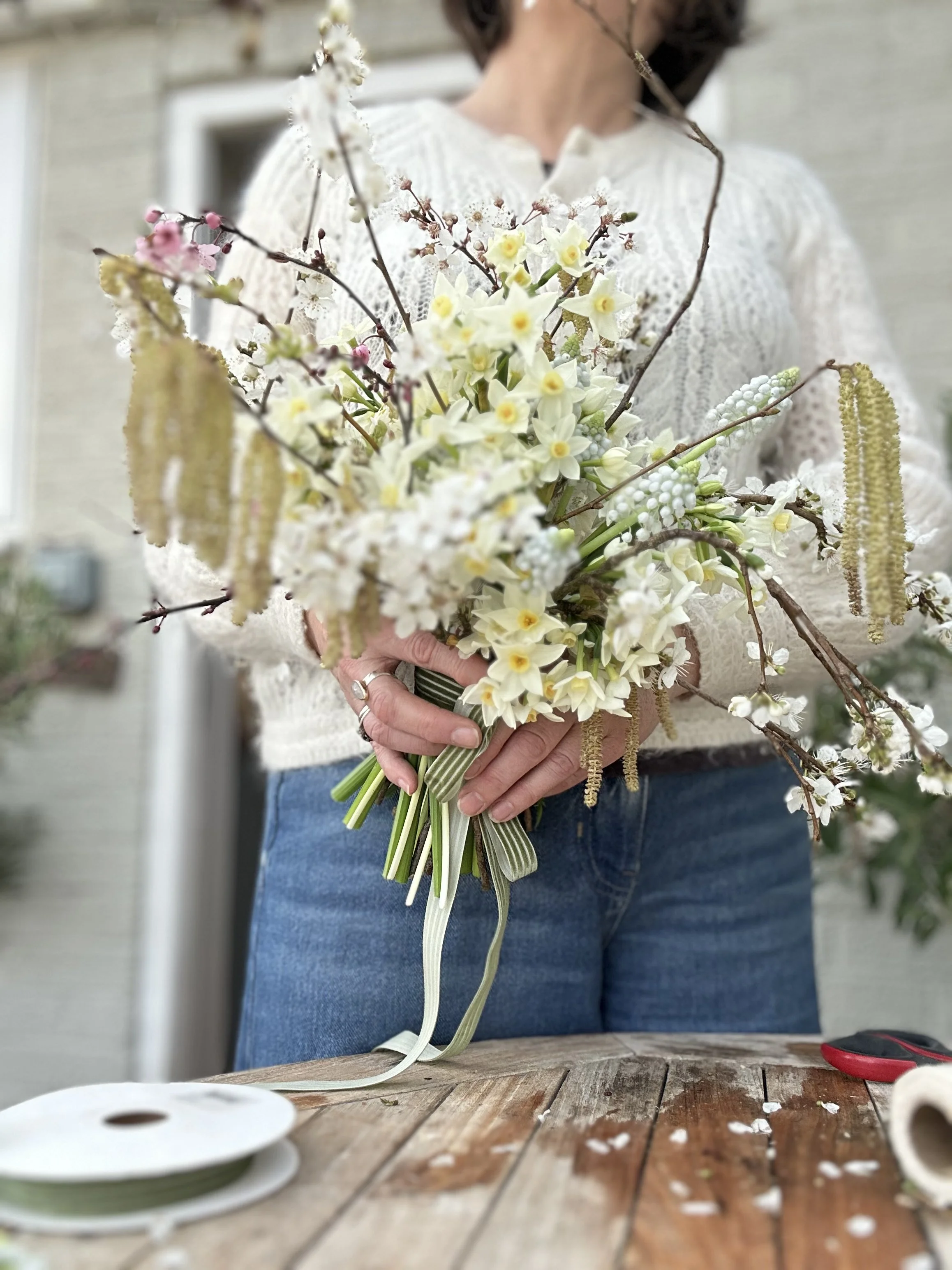 Person holding a bouquet of white and pink flowers, with a blurred background, sitting at a wooden table.