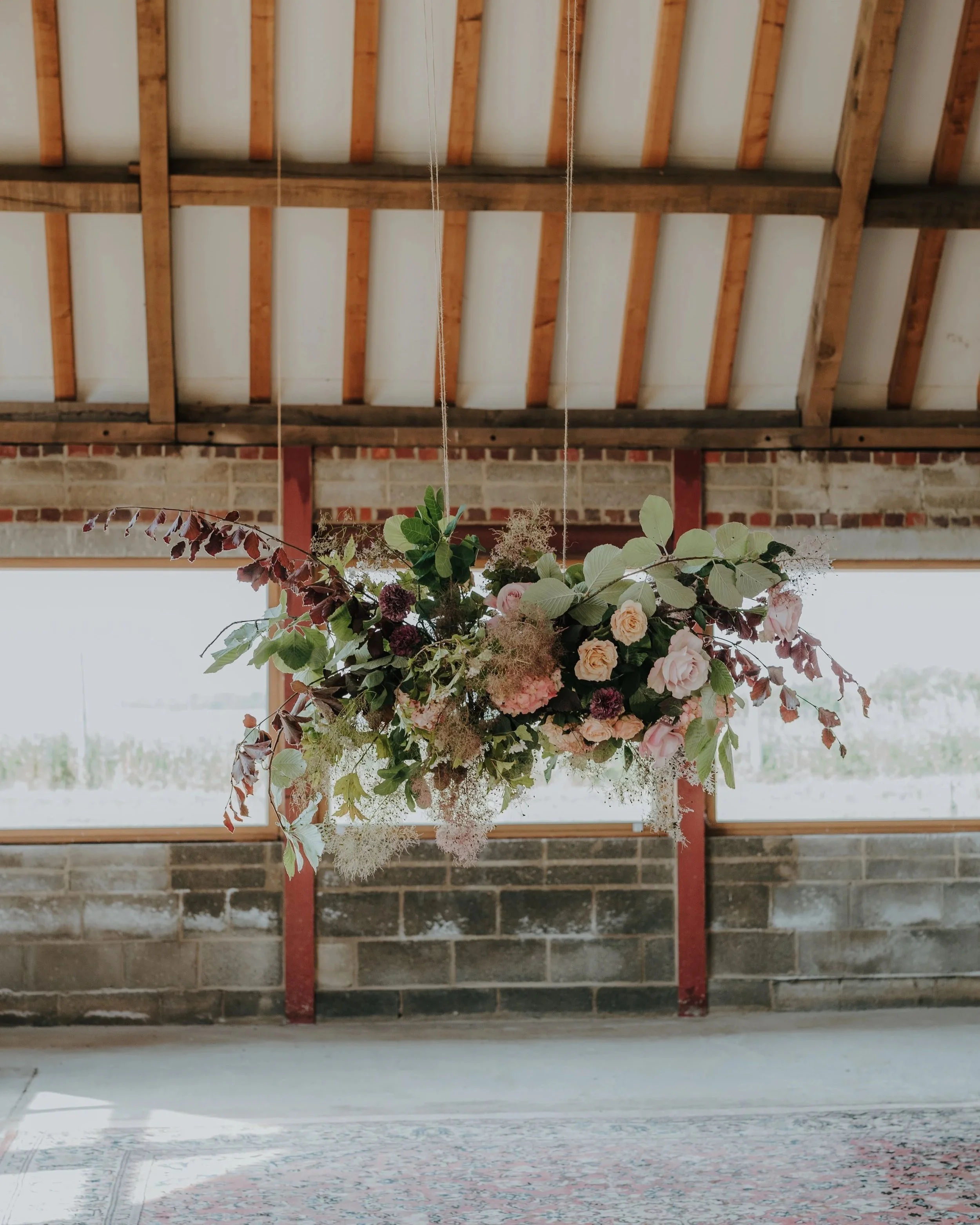 A hanging floral arrangement of assorted pink roses, green leaves, and other flowers in front of a high ceiling with exposed wooden beams and a brick wall.