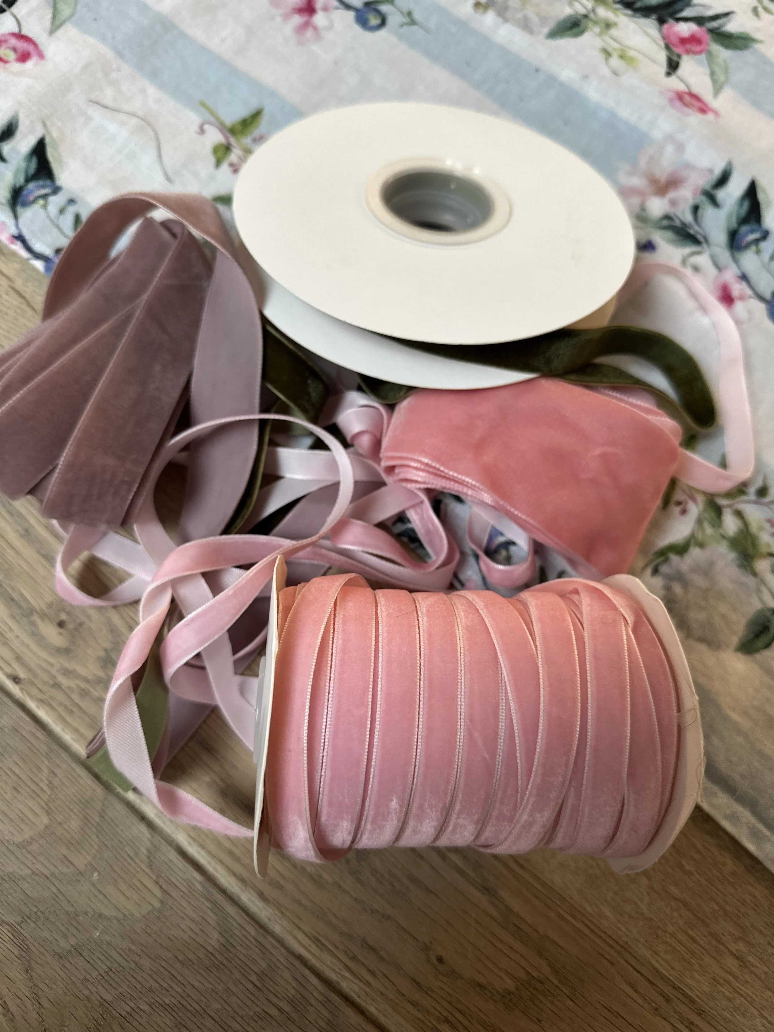 Various rolls of ribbon in pink, peach, and brown shades, some with satin finish, placed on a wooden surface next to a floral tablecloth.