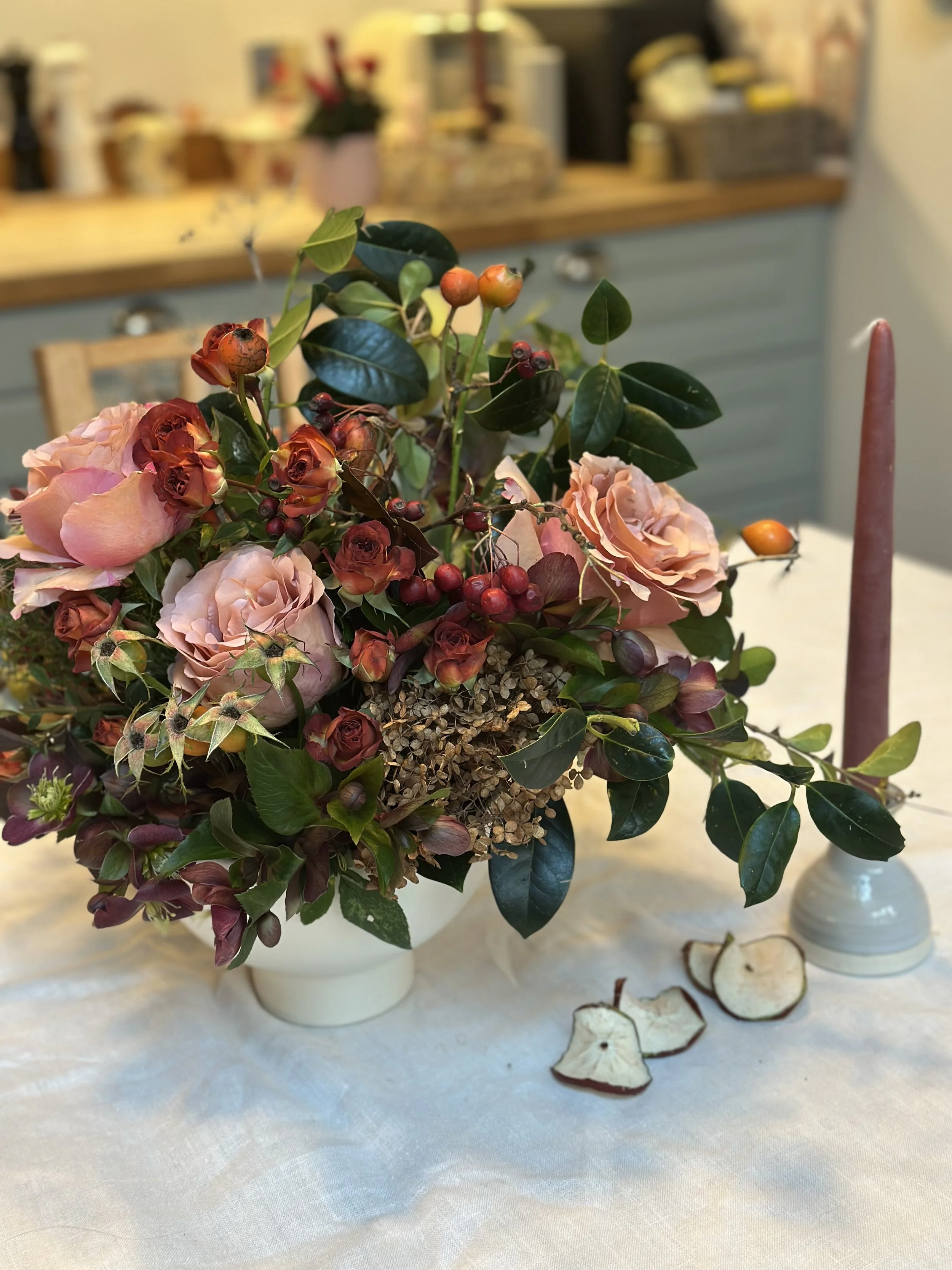 A flower arrangement with pink roses, red berries, green leaves, and dried white flowers in a white vase, placed on a white cloth. Dried apple slices are on the table near the vase.