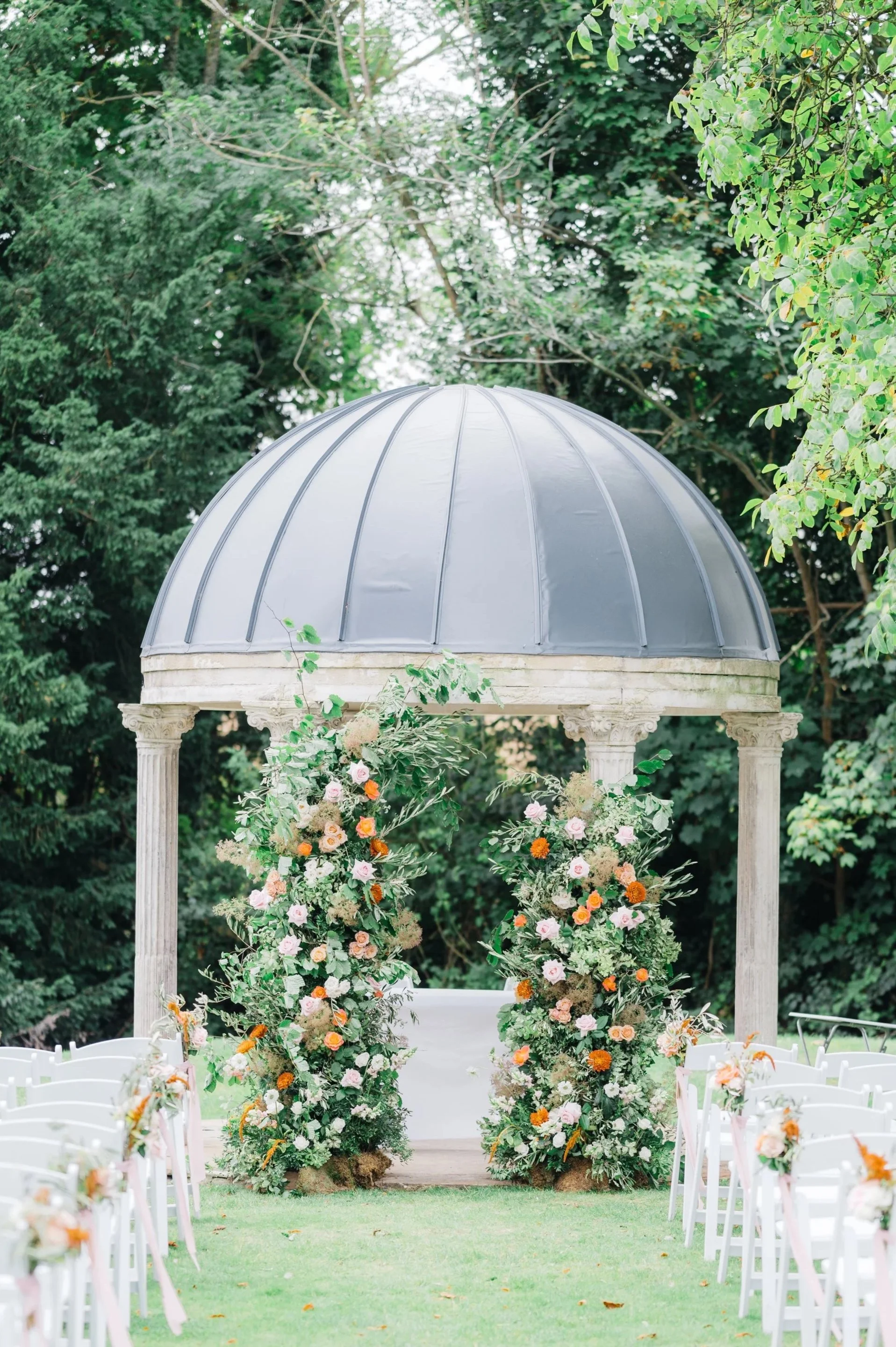 An outdoor wedding ceremony setup with white chairs, floral arrangements, and a classical-style altar under a domed gazebo surrounded by green trees.