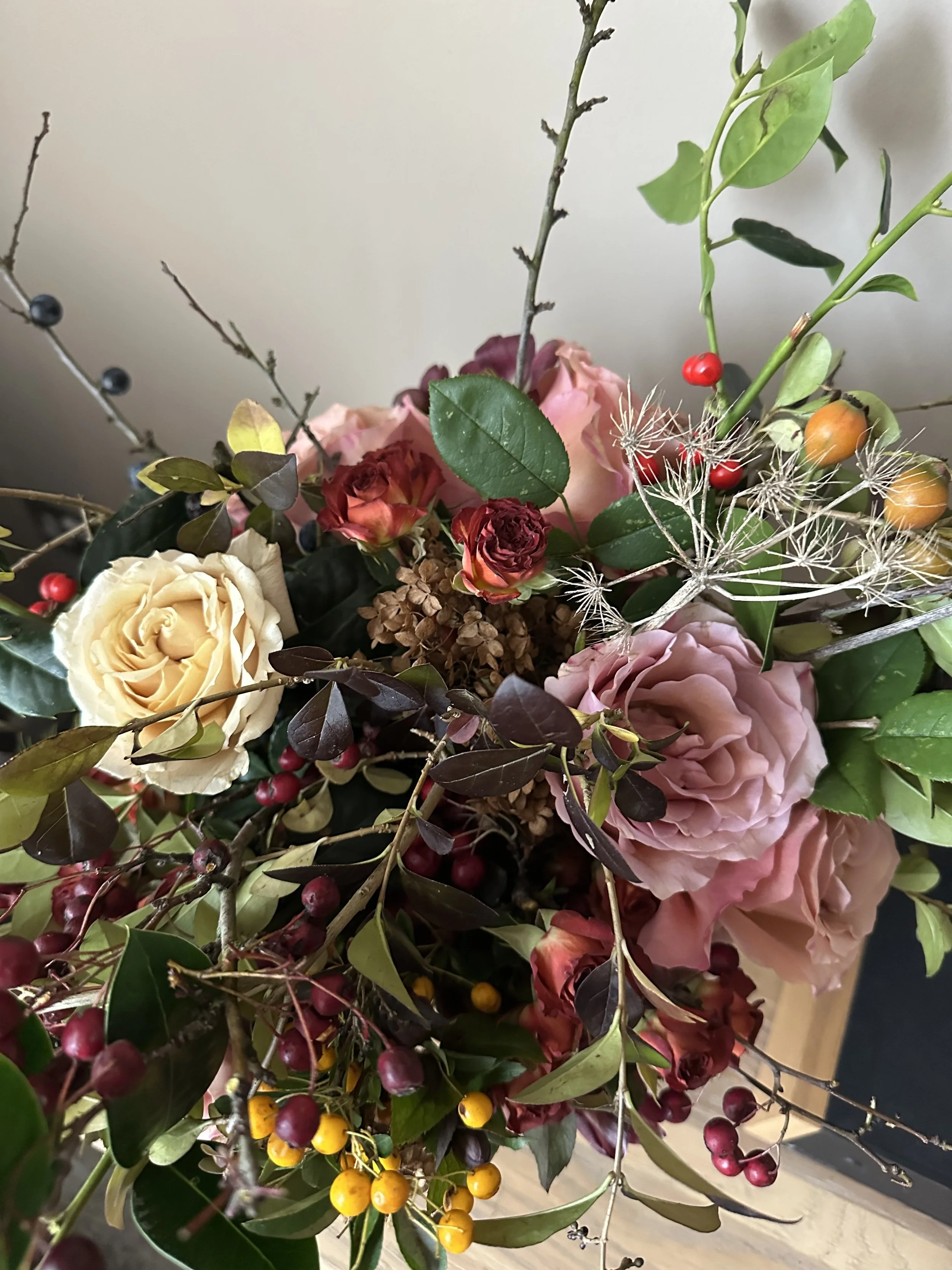 A close-up of a floral arrangement with roses, greenery, berries, and decorative dried flowers.