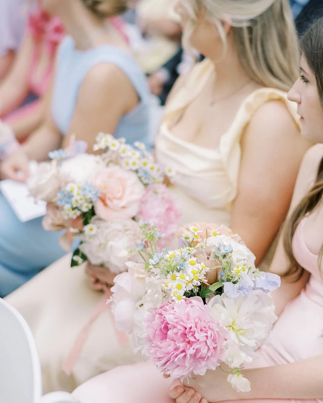 Women dressed in pastel-colored dresses attending a wedding, holding bouquets of pink, white, and yellow flowers.