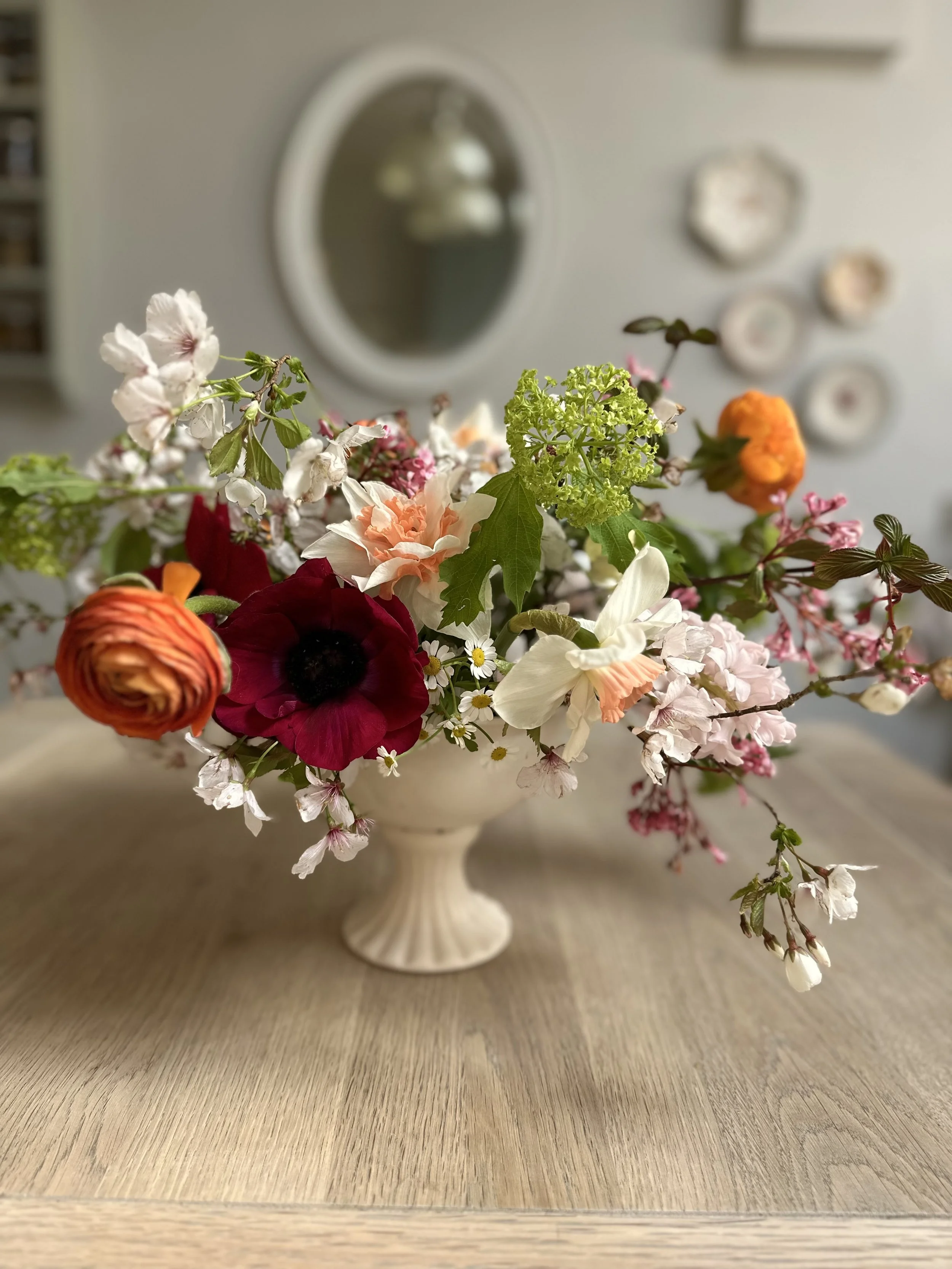 A white ceramic vase with various colorful flowers including white, pink, orange, and dark red blooms, placed on a wooden surface in front of a wall decorated with small plates and a mirror.
