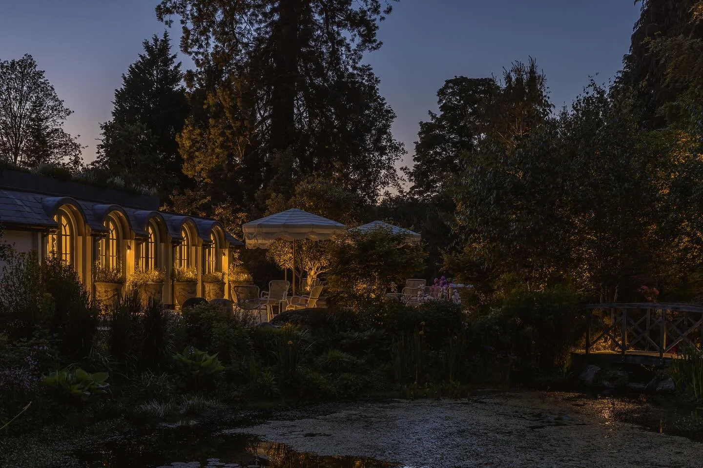 A view across a pond to the decking outside the indoor swimming pool. It&rsquo;s the glow from inside that provides a soft warm light to the seating area outside. As that fades away some low output spike lights highlight some of the planting and tree