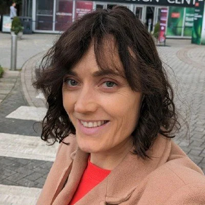 Woman in red top and beige coat with short dark hair smiling in the street