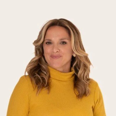 Woman with blonde wavy hair smiling in mustard yellow jumper in front of cream background