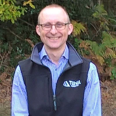 Man in blue shirt stands smiling in front of trees