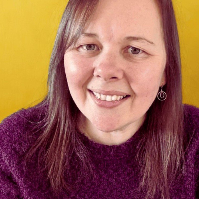 Woman with burgundy jumper and brown straight hair smiling in front of yellow background