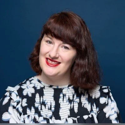 Woman with dark hair and a fringe smiles in front of dark blue background