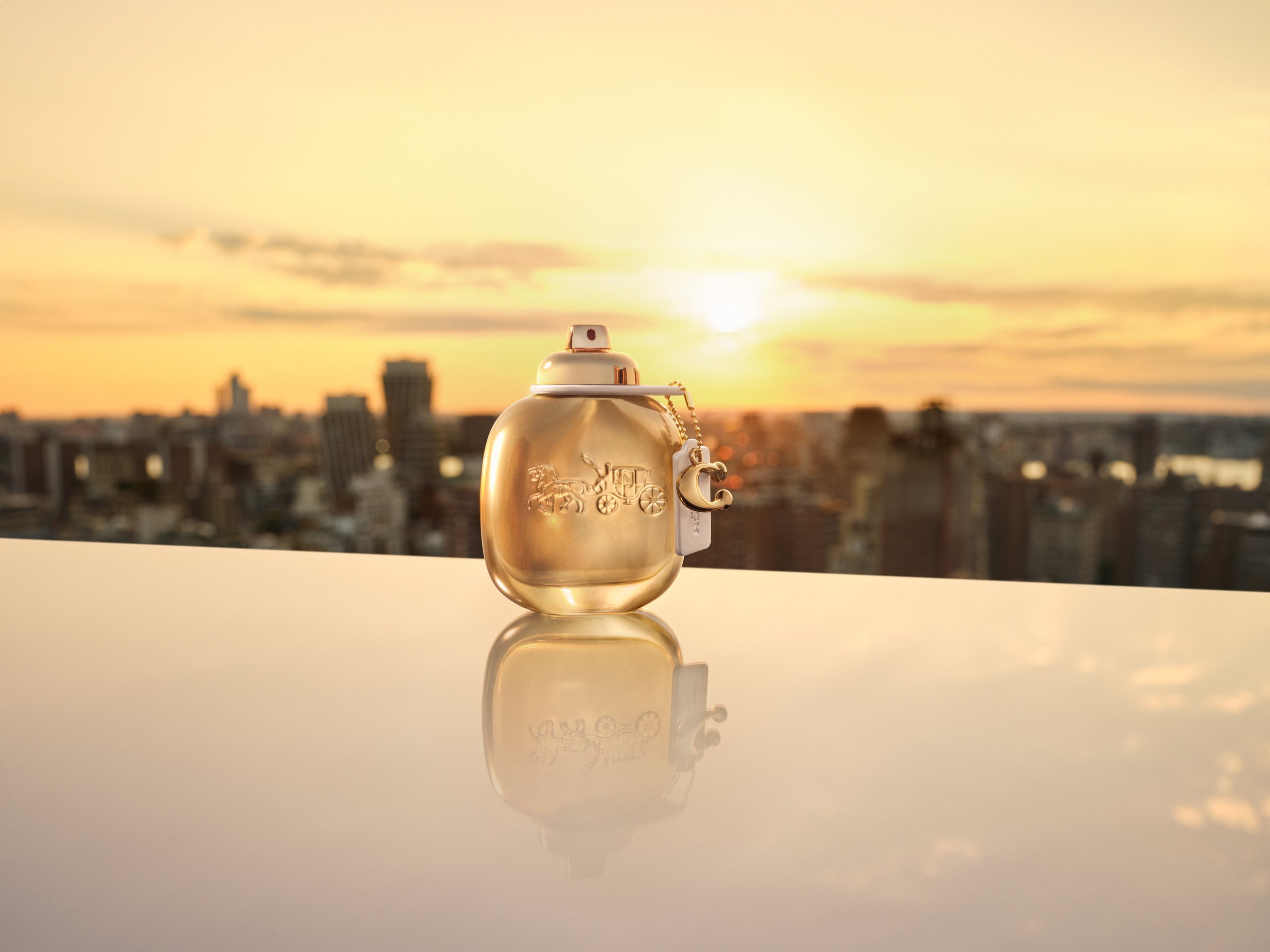 Coach Gold eau de parfum bottle on reflective surface against golden sunset sky, photographed by Cass Bird