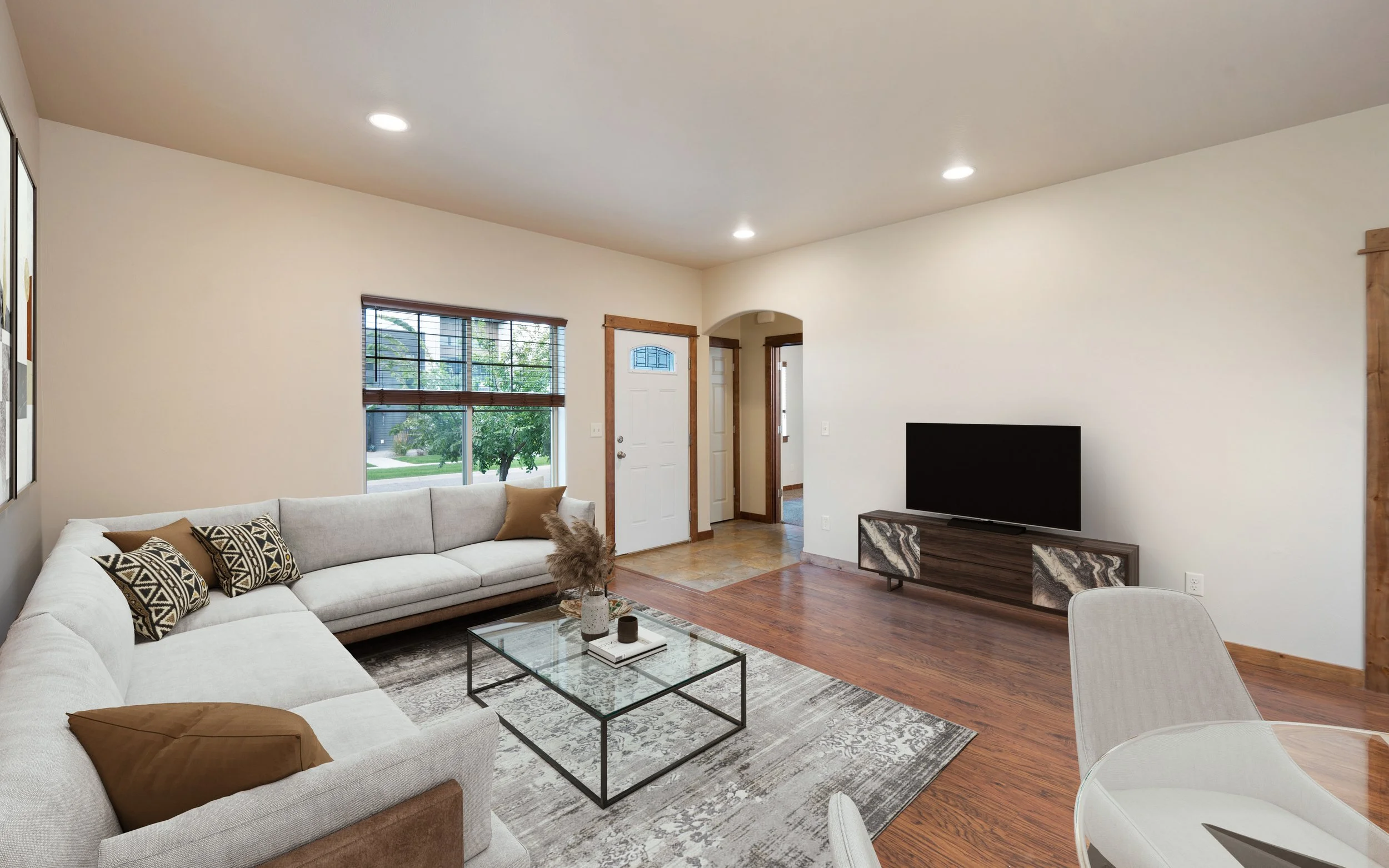 Living room with beige walls and hardwood floors, featuring a large white sectional sofa with patterned and plain throw pillows, a glass coffee table with decorative items, a flat-screen TV on a sleek wooden console, and a window with blinds overlooking a green yard.
