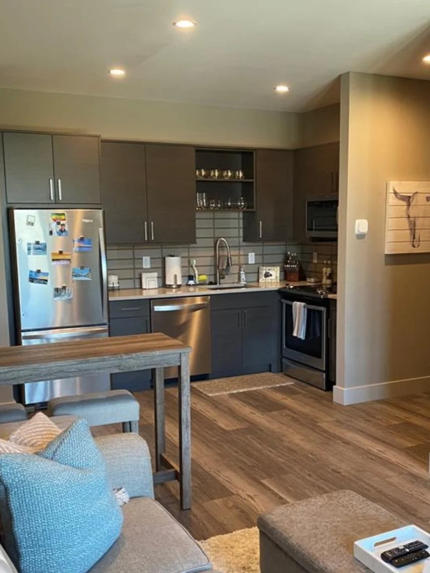 Modern kitchen with grey cabinets, stainless steel refrigerator, dishwasher, and oven. Open shelving with glasses. Small dining area with a wooden table and cushioned chairs, adjacent to the living area with a beige couch and pillows.