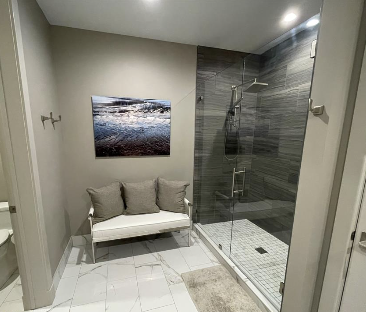 A small modern bathroom featuring a glass-enclosed shower with gray tile walls and white square tiles on the shower floor, a beige rug on a white marble floor, a white bench with gray cushions, a cactus-shaped wall hook, a piece of wall art resembling ice or snow, and part of a toilet visible in a nearby room.