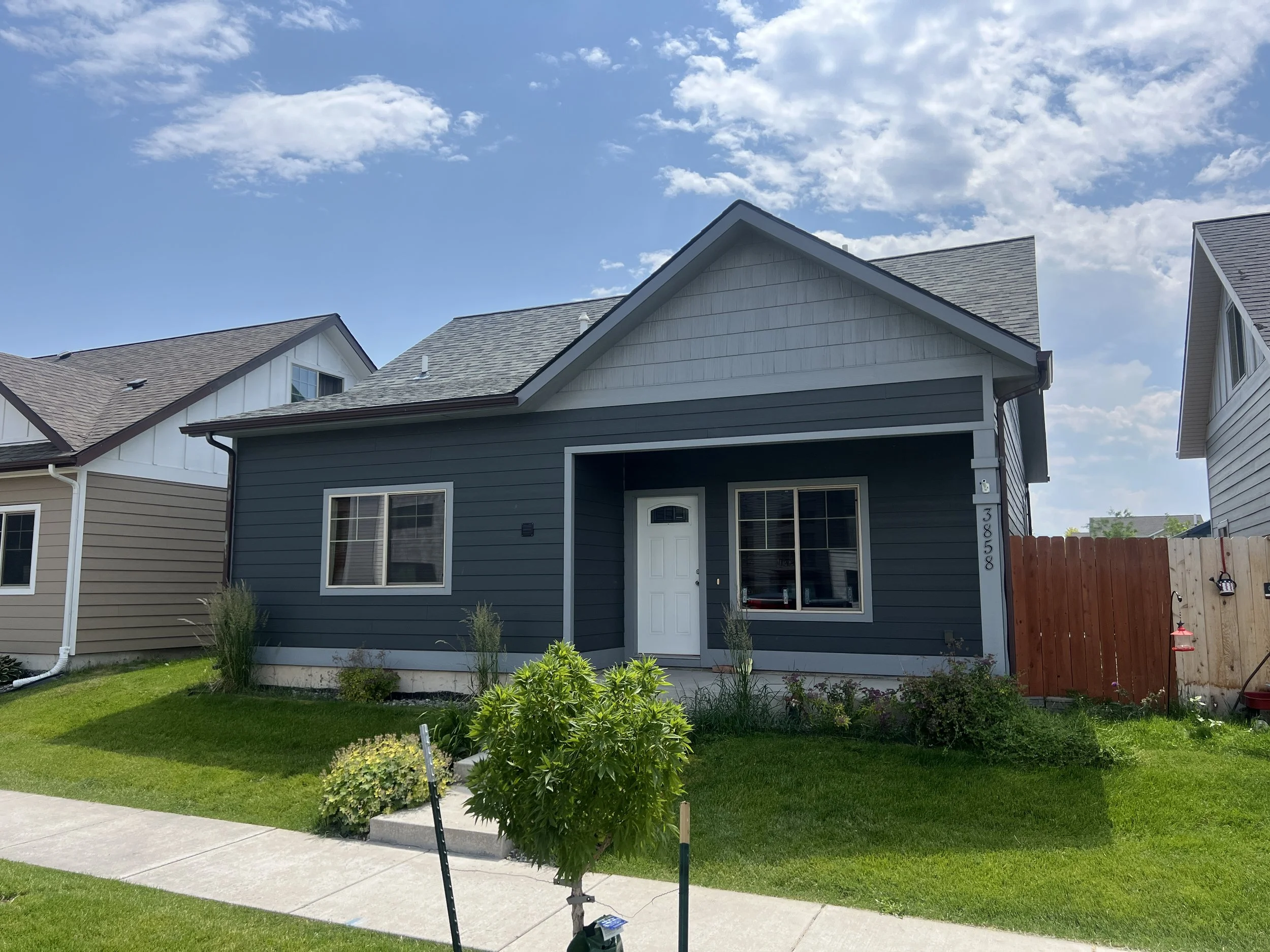 Front view of a modern two-story house with dark blue siding, white trim, and a gray roof, surrounded by a well-maintained lawn and neighboring houses under a blue sky with scattered clouds.