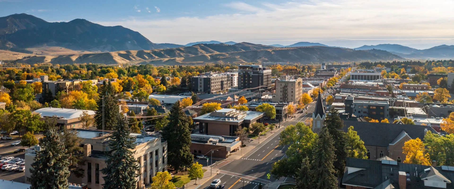 A panoramic view of a small town with colorful autumn trees, tall buildings, a church with a steeple, and mountains in the background under a clear sky.
