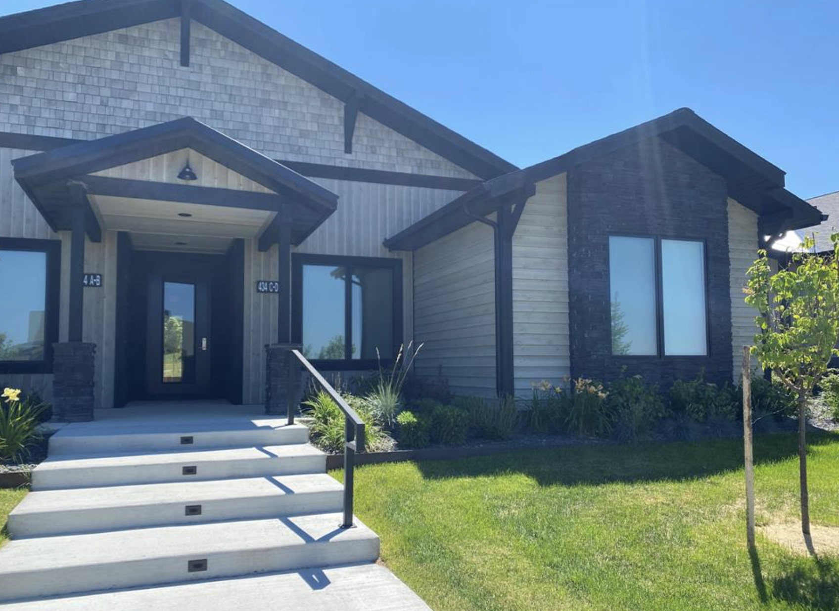 New modern house with a front porch, stairs, and lush green yard with small trees and plants.
