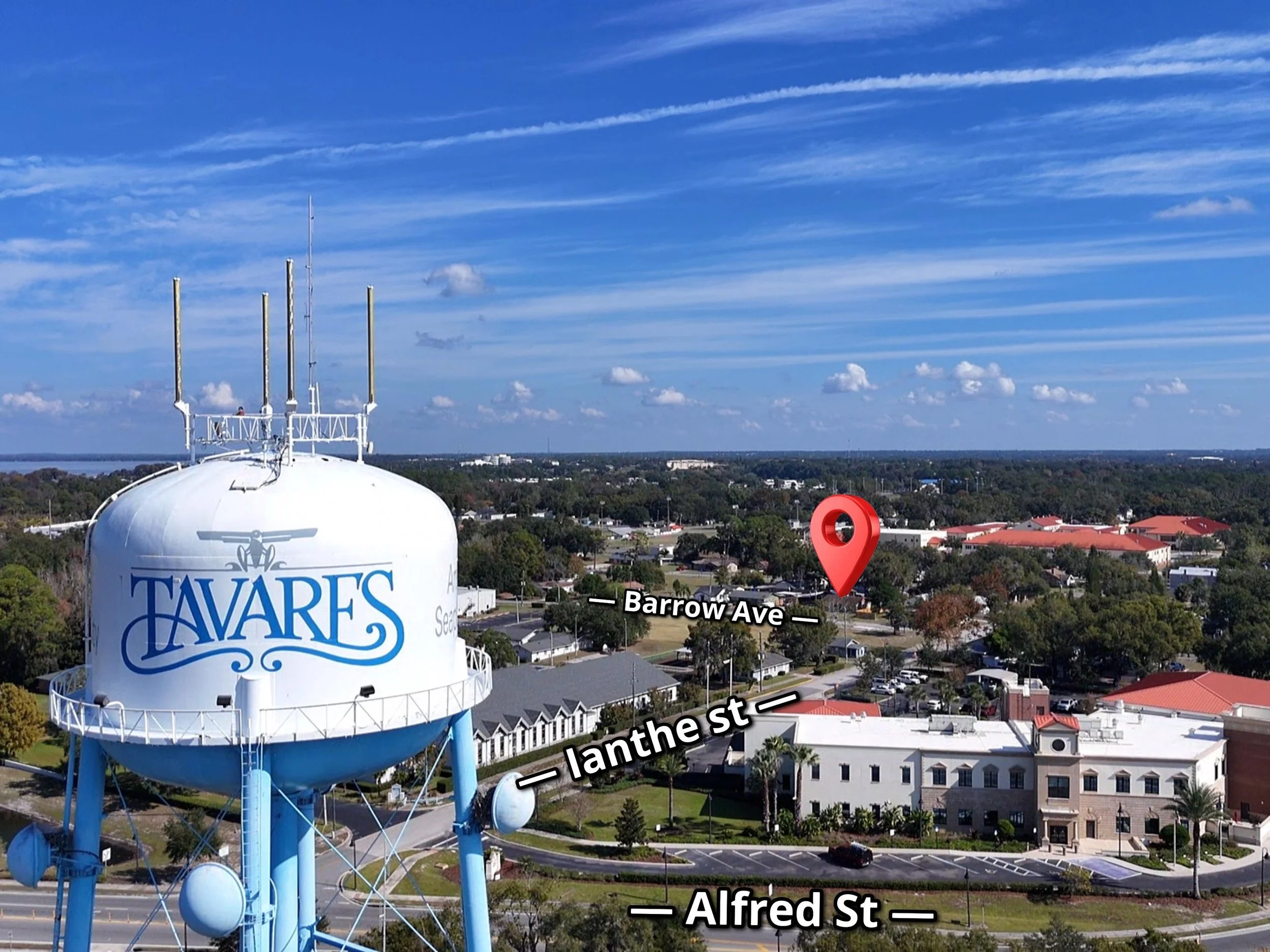 A large white water tower with the words 'Tavares' written on it, and a logo of an angel holding a trumpet. The water tower is situated in a cityscape with trees, buildings, and a parking lot. The sky is mostly clear with a few clouds and contrails.