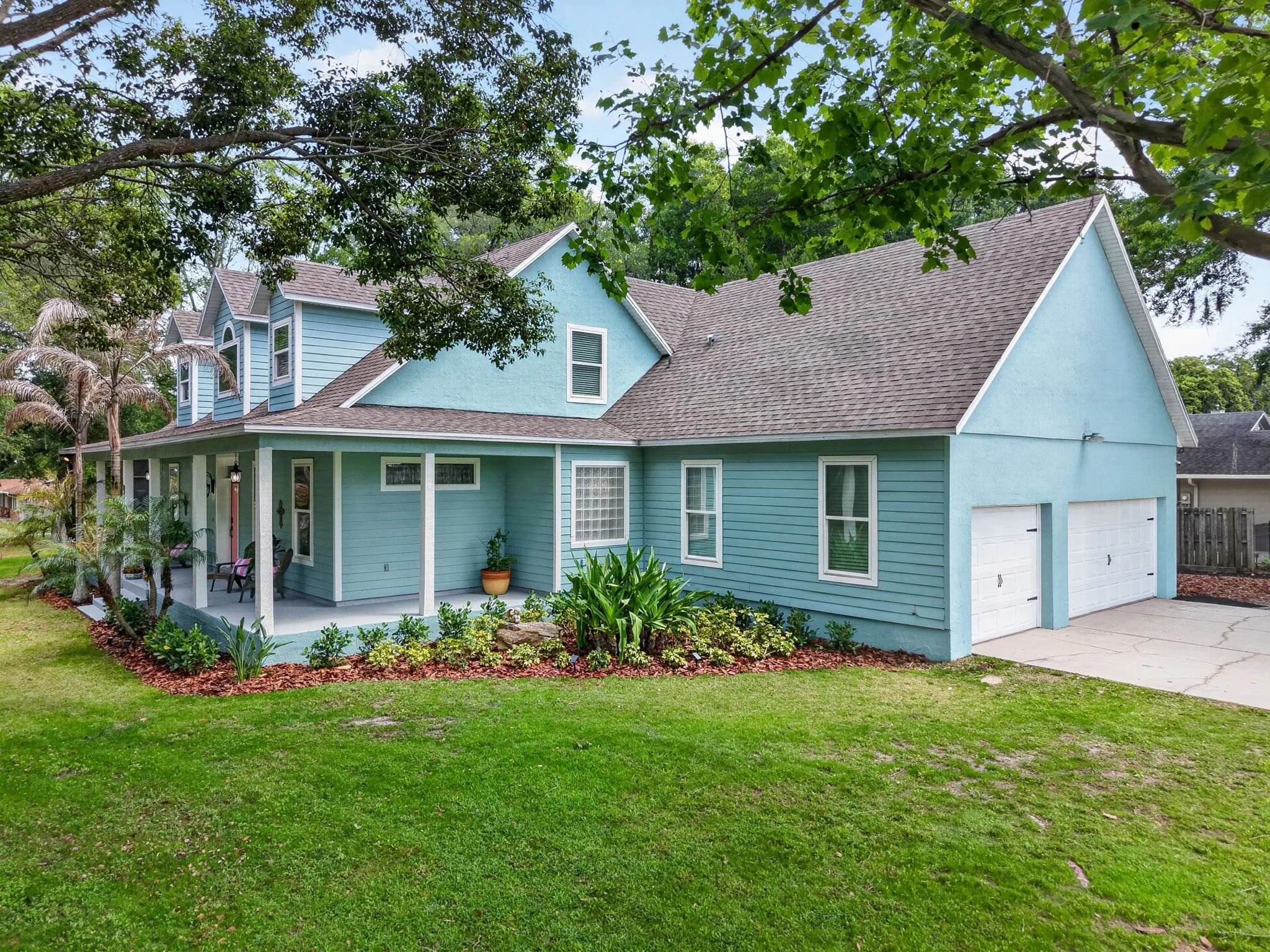 A light blue two-story house with a gable roof, white trim, and a front porch, surrounded by green grass and trees.