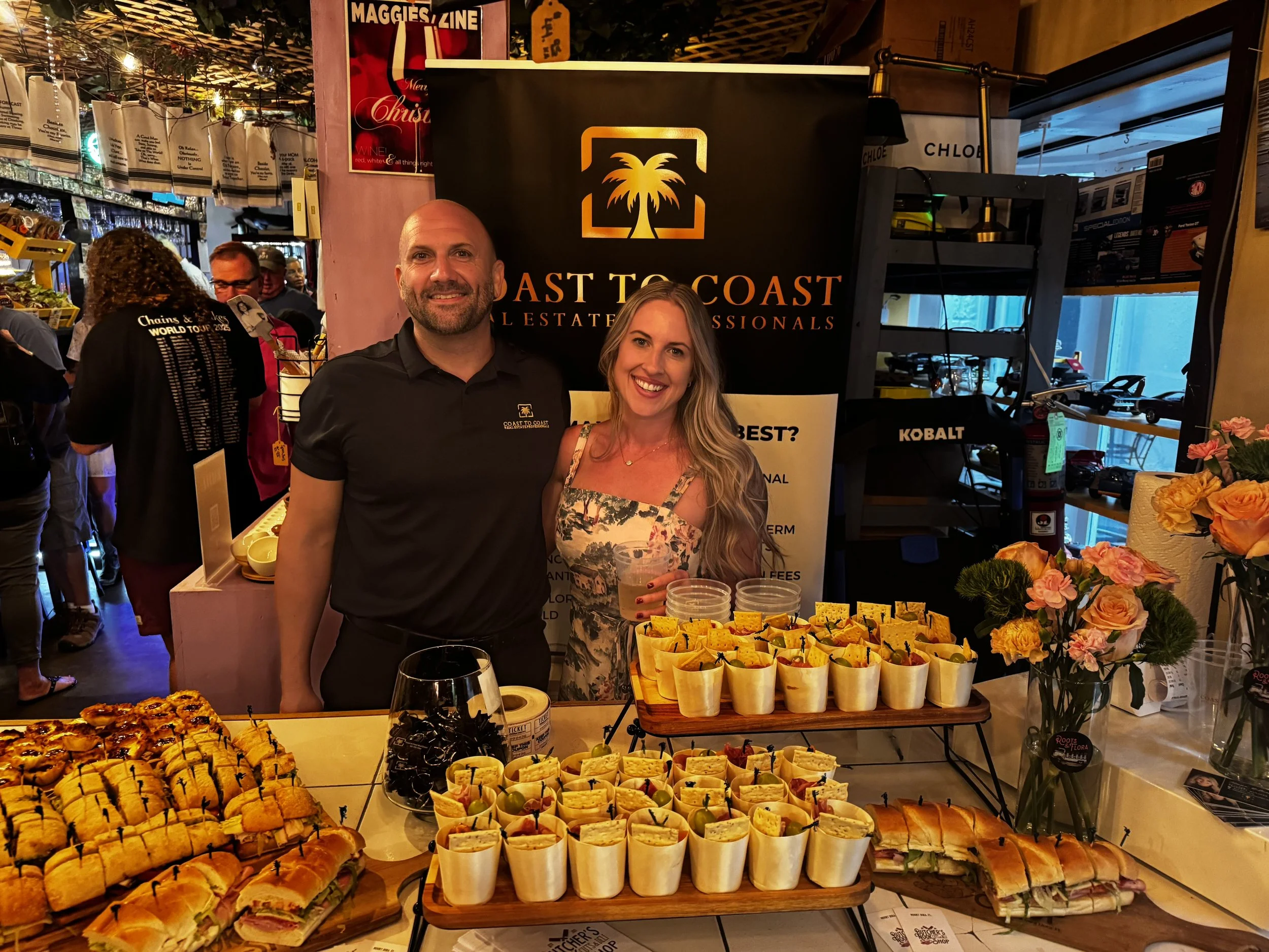 Two people stand behind a table with an assortment of small sandwiches and snacks, in front of a black sign with gold palm tree logo and text, at a social event or market.