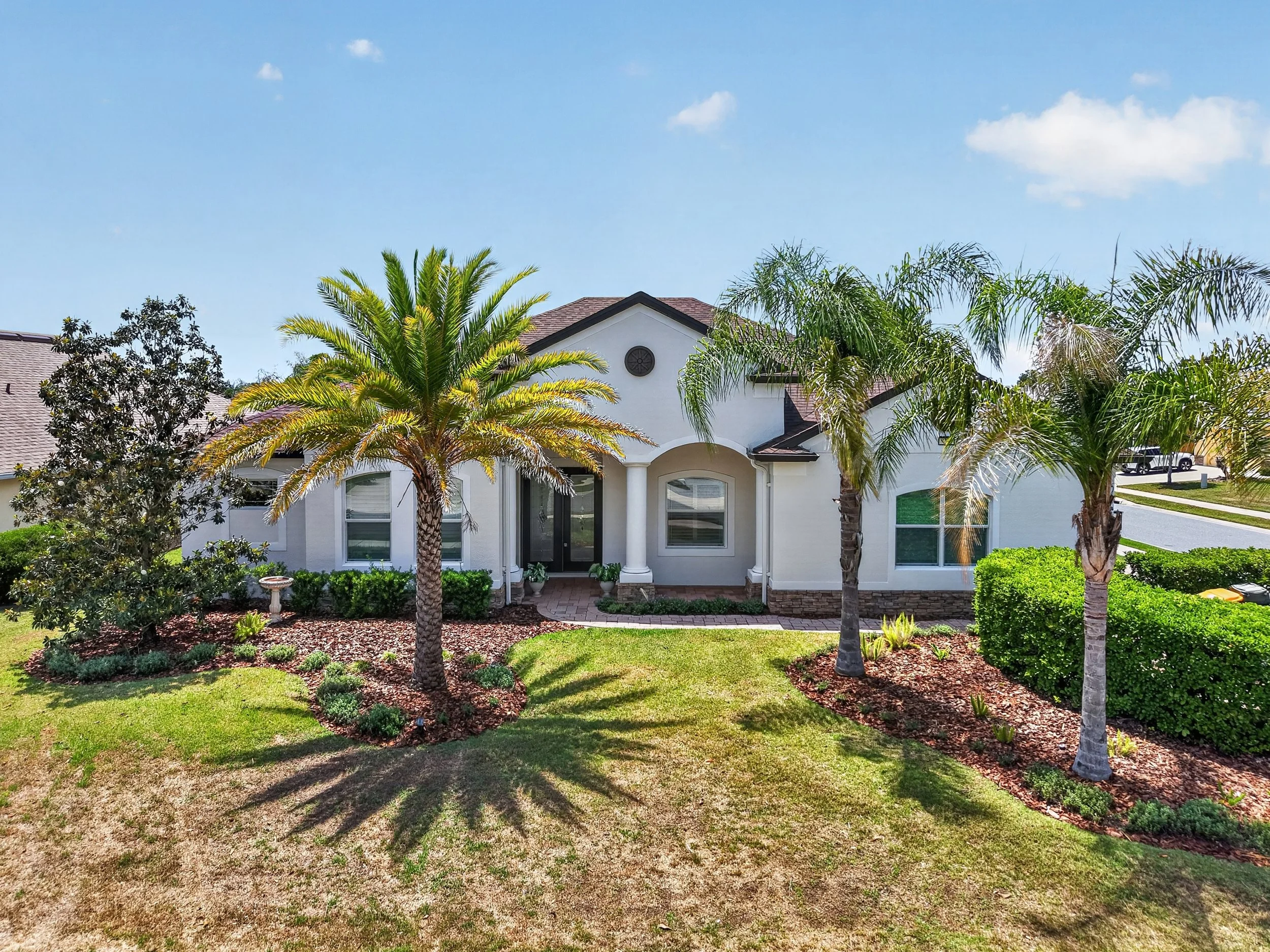 A white house with a dark roof surrounded by palm trees and a well-maintained lawn under a blue sky with a few clouds.