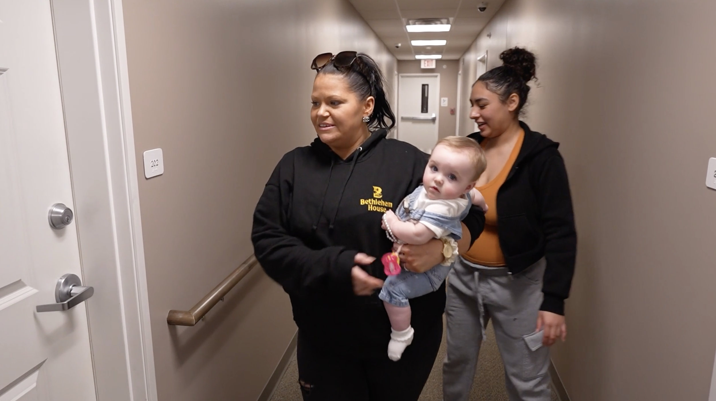 Two women walking down a hospital hallway with a baby. One woman has dark hair tied up, wears sunglasses on her head, and a black hoodie; the other has curly hair in a bun, wears a black hoodie and an orange top. The woman holding the baby is wearing a black hoodie with yellow text that reads 'Bethlehem House.' The baby has light hair, is dressed in a white and blue outfit, and is holding a pink toy. The hallway has beige walls, white doors, and a handrail.