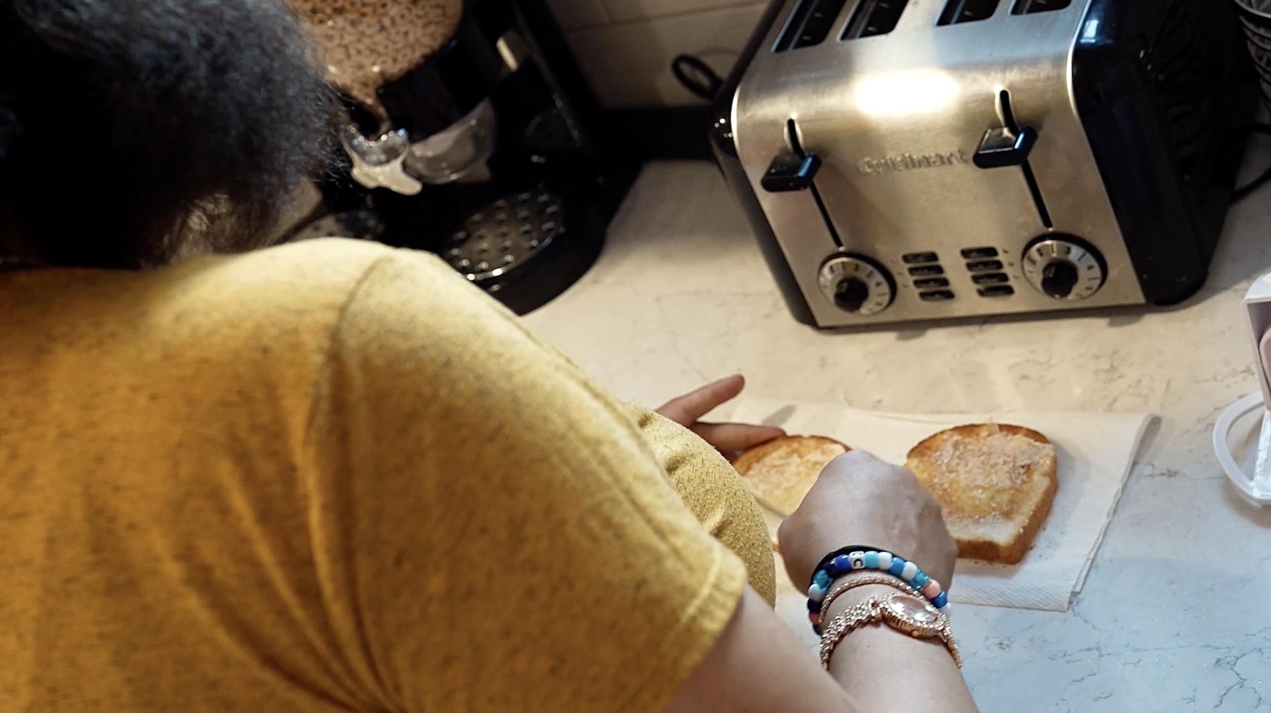 A person wearing a yellow shirt toasting bread slices on a paper towel in a kitchen with a toaster and coffee maker in the background.