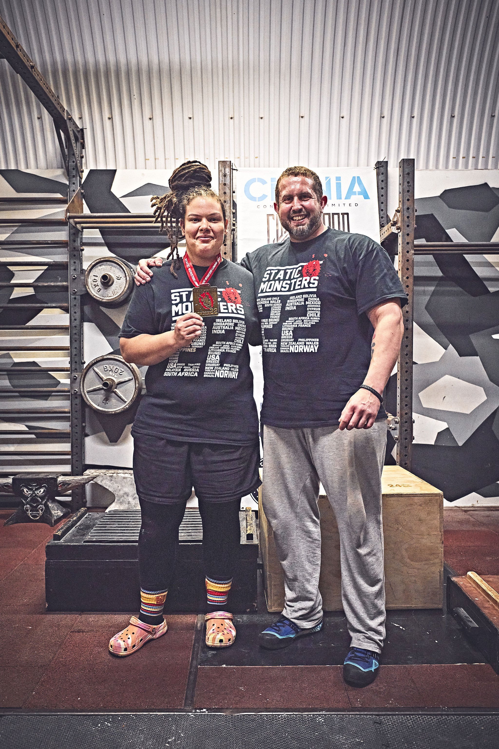 Two people standing in a gym, one with dreadlocks holding a medal and wearing colorful sandals and striped socks, both wearing black "Static Monsters" T-shirts, smiling at the camera. The background includes gym equipment and a white banner.