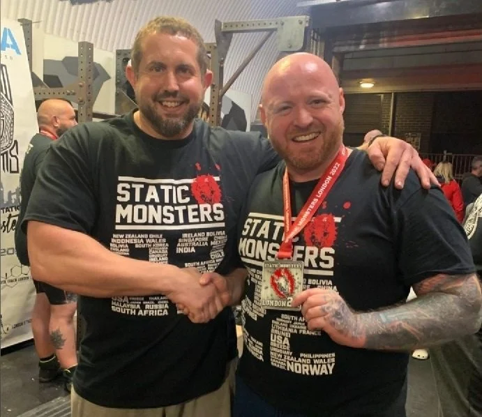 Two smiling men wearing black T-shirts with 'STATIC MONSTERS' written on them, shaking hands at an indoor event. One man is holding a medal, and the background includes other people and gym equipment.