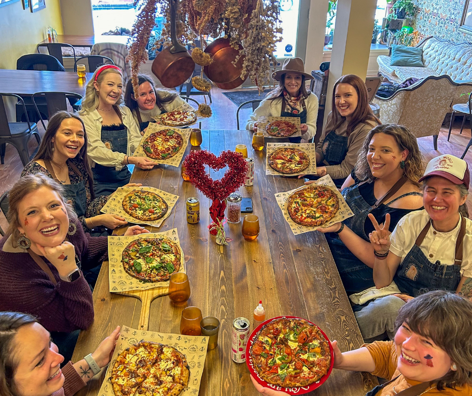 Group of women sitting around a table celebrating with pizzas, drinks, and decorations, including a red heart-shaped centerpiece, in a cozy, decorated indoor setting.