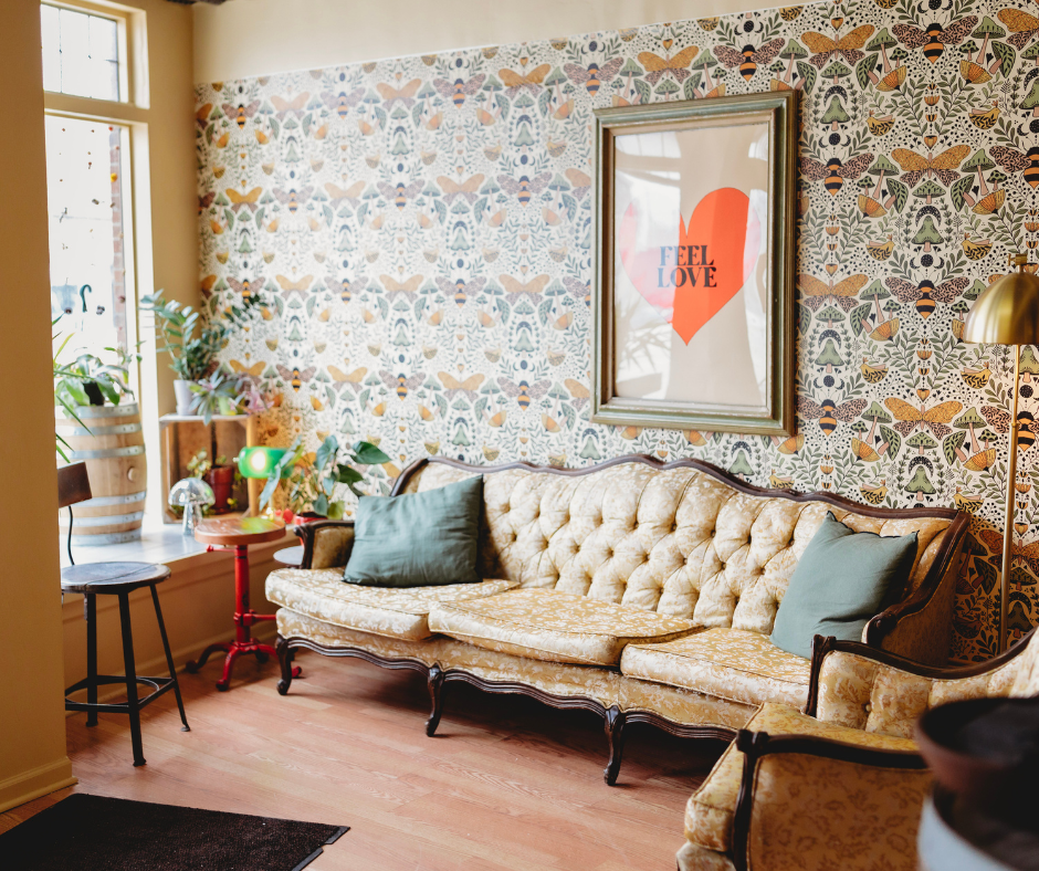 A cozy living room with a vintage floral-patterned wallpaper, an ornate beige tufted sofa with two green pillows, a wooden armchair, a side table with plants, a framed art piece with a red heart and 'FEEL LOVE' text, and sunlight coming through the window.