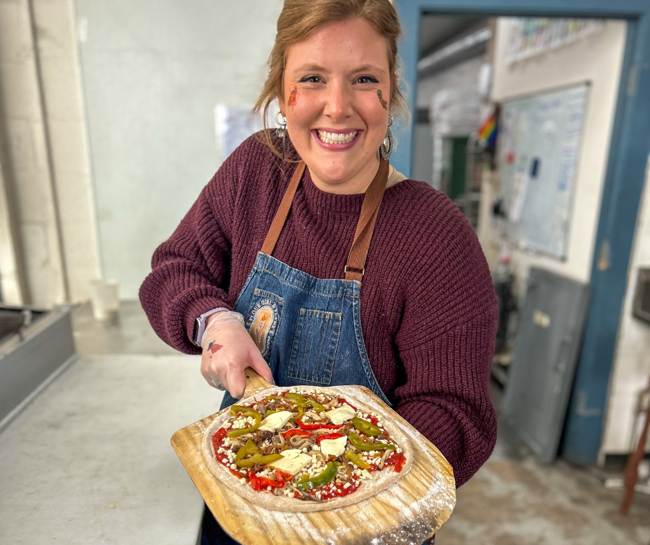 A woman smiling and holding a pizza with peppers and cheese in a kitchen or bakery setting.