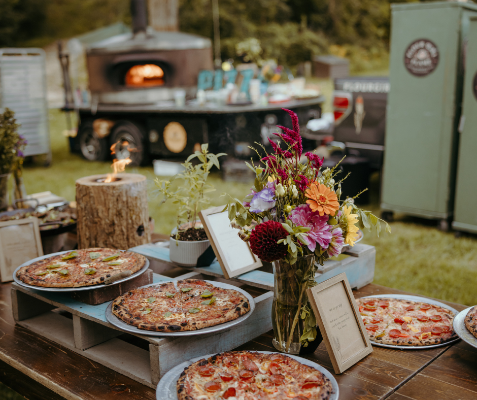 Several pizzas on plates and a pizza cutter on a rustic wooden table, with a large vase of colorful flowers and a potted plant, at an outdoor gathering.