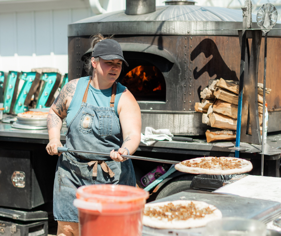 Outdoor pizza food truck with a wood-fired oven, illuminated 'PIZZA' sign, and a table with metal cups against a backdrop of trees and greenery.