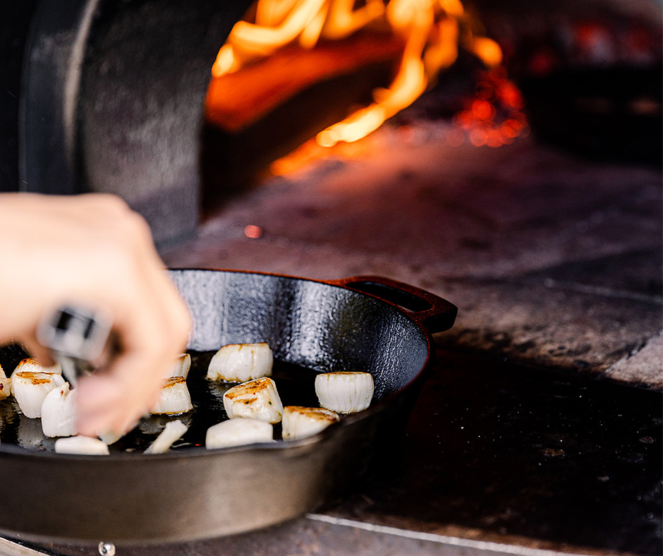 People serving themselves food at an outdoor gathering with flowers and a log with a flame on top in the center.