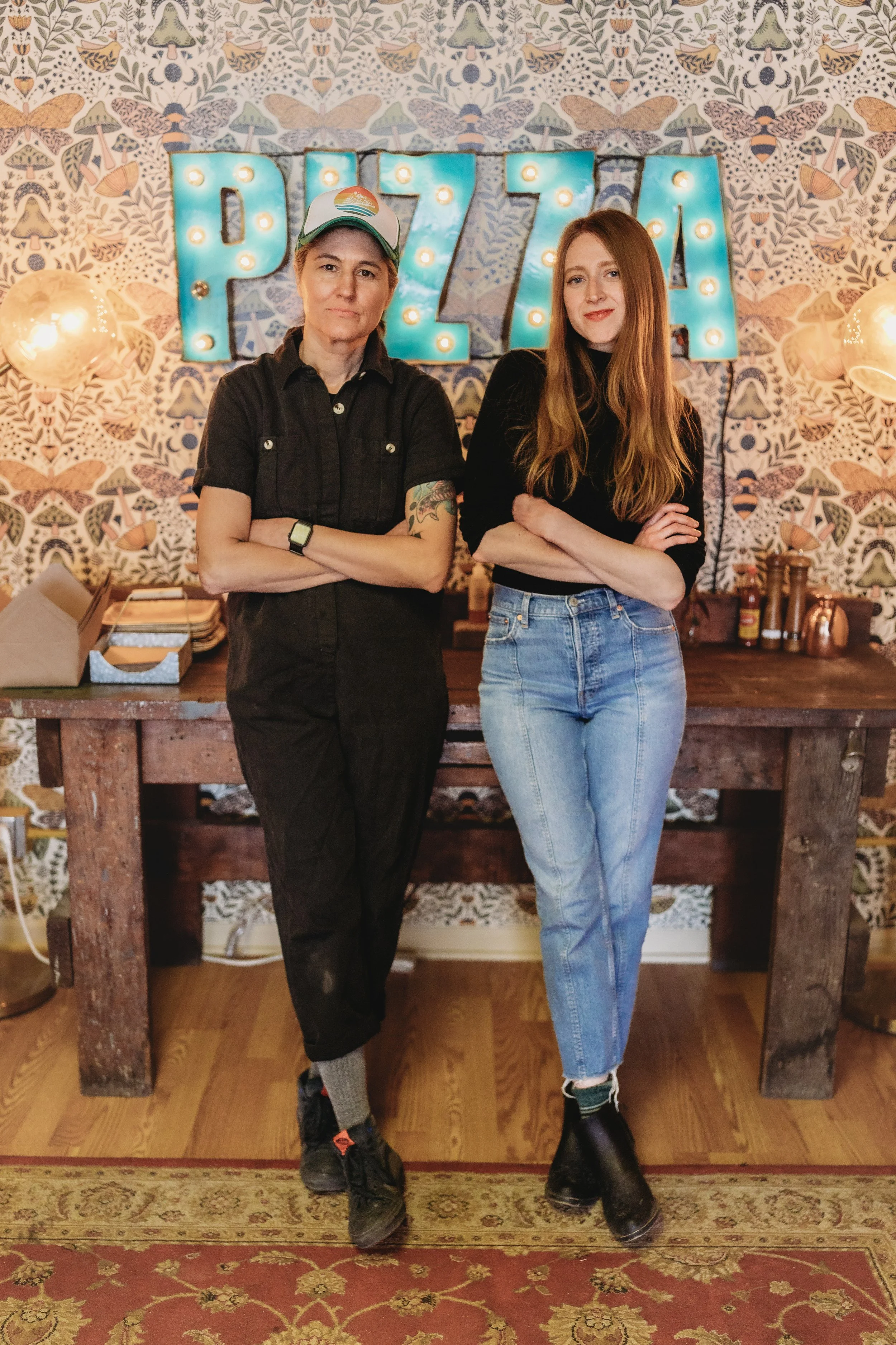 Two women standing with arms crossed in front of a vintage-style wallpaper with large illuminated letter sign spelling 'PIZZA' in blue lights. One woman has short hair and tattoos, and the other has long red hair. There is a wooden table with condiments behind them.