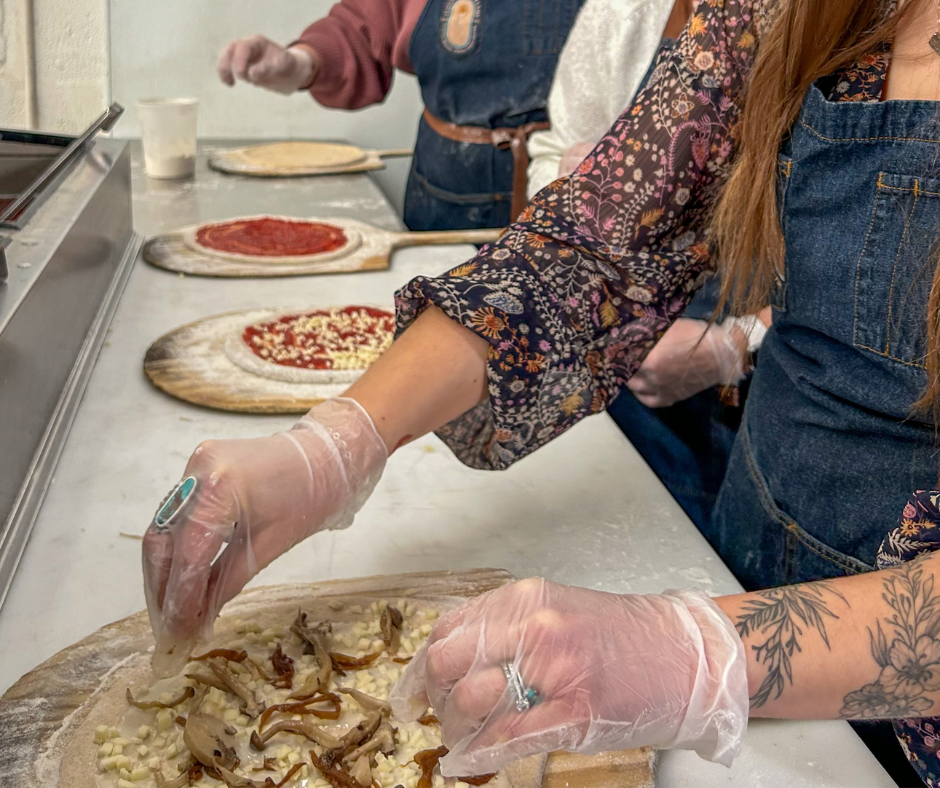 People preparing to make pizza with toppings like mushrooms and cheese in a kitchen.