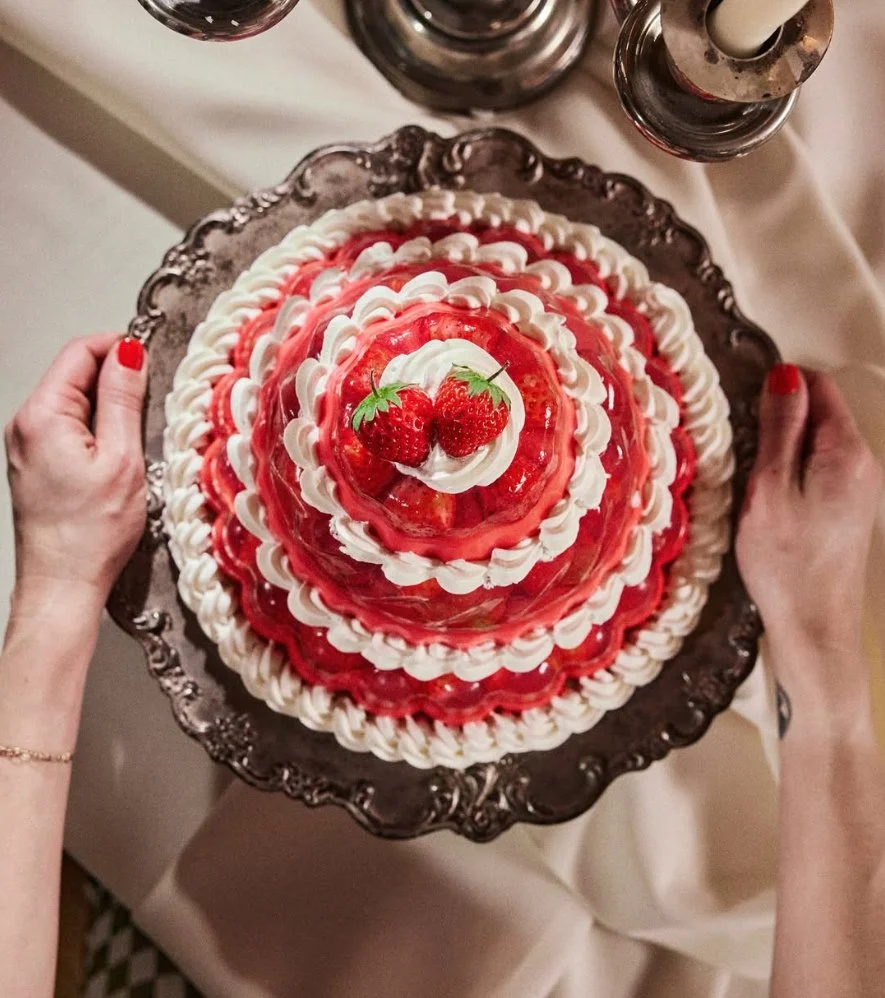A layered cake with white and red frosting, topped with two strawberries and garnished with edible greenery, placed on a decorative silver platter.